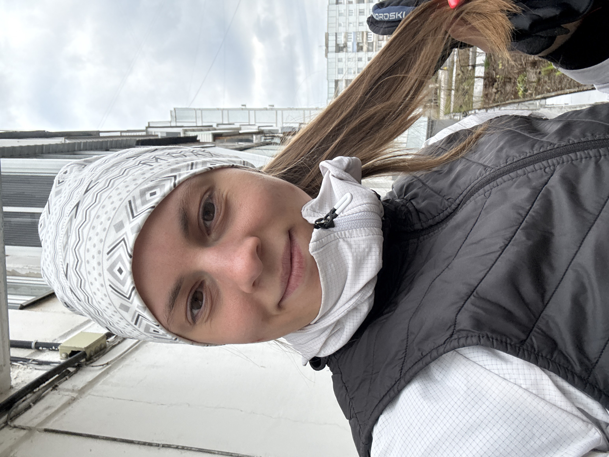 This angled outdoor portrait captures a young woman in Chaikovsky, Russia, under an overcast sky. She is smiling faintly and looking slightly upwards. Her medium-brown hair flows past her shoulder, and she holds a section of it with her right hand, which is gloved. The black glove has a blue and white band with the text "NORD SKI" visible. She wears a grey and white patterned headband, a white long-sleeved athletic top with a subtle grid texture, and a black padded vest. A white neck gaiter is also visible around her neck. In the background, urban buildings stand beneath a cloudy sky, with hints of distant trees or foliage to her right. The foreground, positioned below her in the image, reveals electrical wires and a junction box on a ledge, suggesting the photo was taken on a balcony or rooftop. The scene conveys a calm, casual outdoor moment.