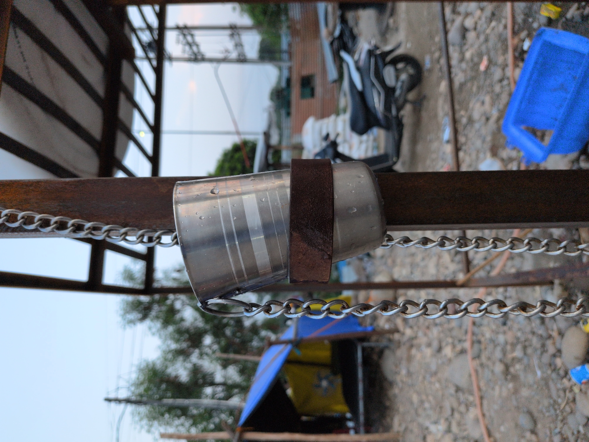 In Jammu, India, two stainless steel cups hang from a metal pole by a chain. The cups have a textured surface with horizontal lines and are wet, indicating they may have been recently used or washed. A rusty metal band encircles the upper cup, securing it to the pole. The background is somewhat blurred, showing a makeshift shelter with blue and yellow tarps, a motorbike parked on a gravelly ground, and a blue plastic container. The overall scene suggests an outdoor, possibly temporary or utilitarian setting in a part of Jammu.