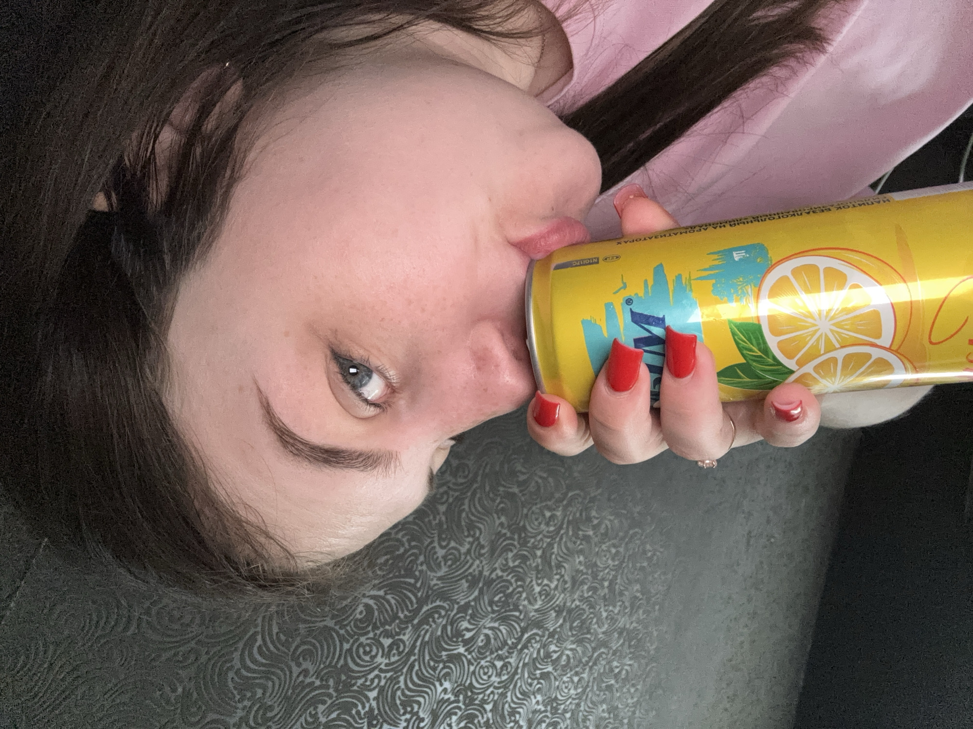 A woman with fair skin, dark brown hair, and red nails is shown drinking from a yellow can with an image of lemons and palm trees, in a horizontal view. She is wearing a pink top, and the background behind her head is a dark gray patterned material. The exact location in Chornomorskoe, Ukraine, cannot be determined without additional information.