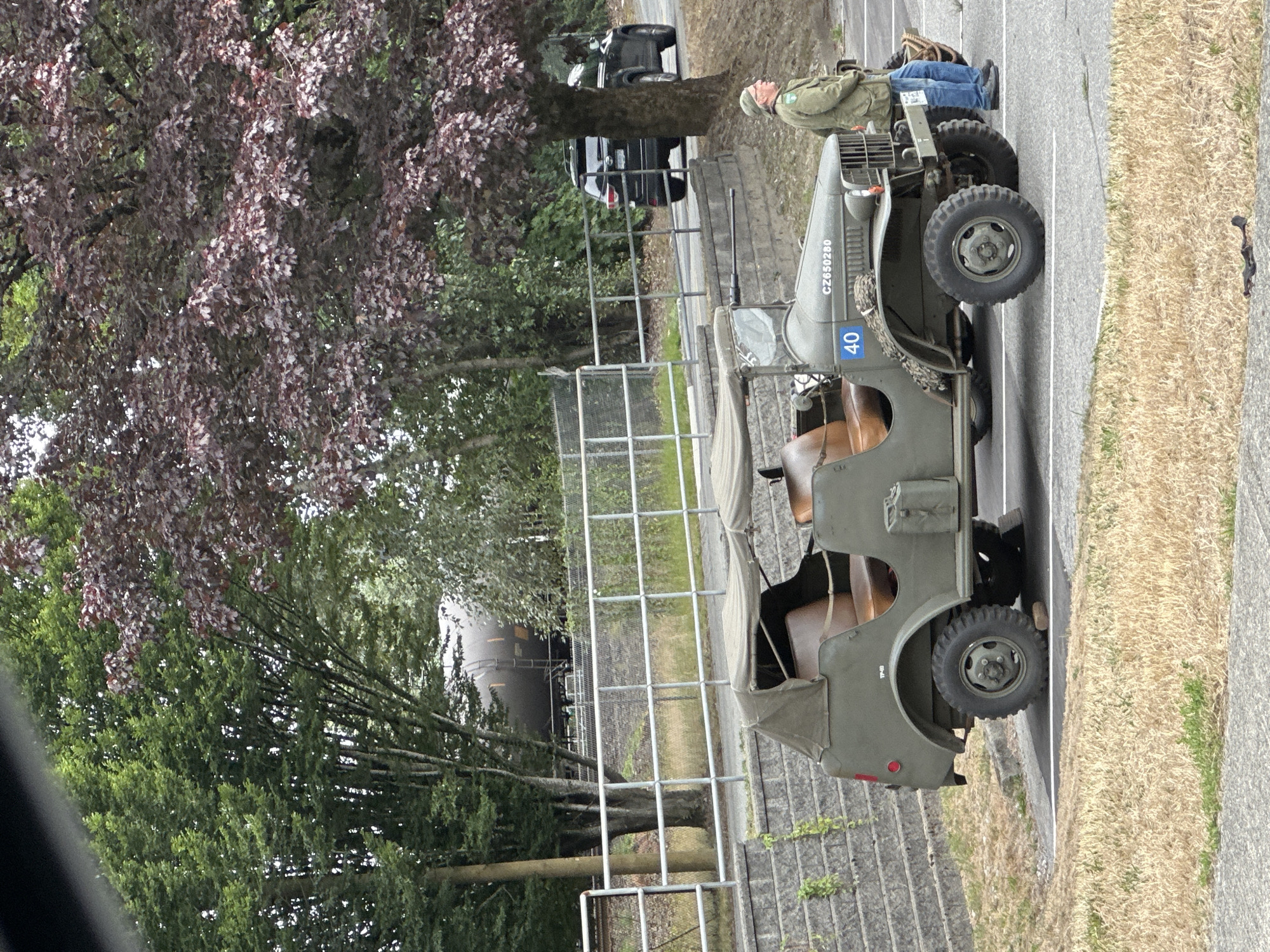 An olive-drab military jeep, identified by the markings "CZ650280" and "40" on its hood, is parked on an asphalt surface, adjacent to a retaining wall constructed of grey concrete blocks. The jeep has a canvas top and tan upholstered seats. A man wearing a green military-style jacket, a green cap, and blue jeans stands beside the jeep, facing to the right of the frame. In the background, a black SUV is parked on a road, and beyond that, a train car is visible near a chain-link fence. Lush green trees and a tree with dark red leaves frame the scene. The ground is covered with dry, yellow grass and patches of green vegetation. The scene is set in Port Coquitlam, Canada.