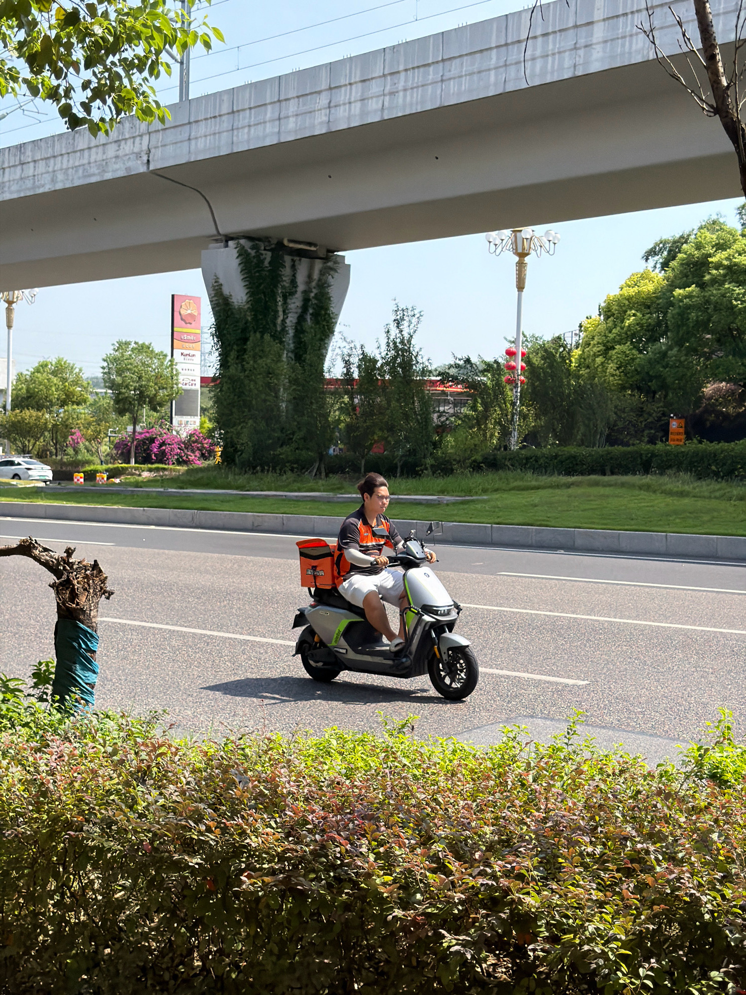 A man rides a grey electric scooter with an orange delivery box attached to the back. He is wearing a grey shirt with orange accents and white shorts. The scooter is on a paved road with white lane markings, indicating it is a city street. In the background, there is a large overpass, trees, and a sign that reads "USmile" and "KunLun". The scene is outdoors on a sunny day, with a clear blue sky. The image is taken from a slightly elevated position, looking down on the road and the scooter. The lower part of the image is filled with green bushes. The overall environment suggests an urban setting, possibly in Jiangjin, China, given the presence of a KunLun sign, which is a well-known gas station brand in China.