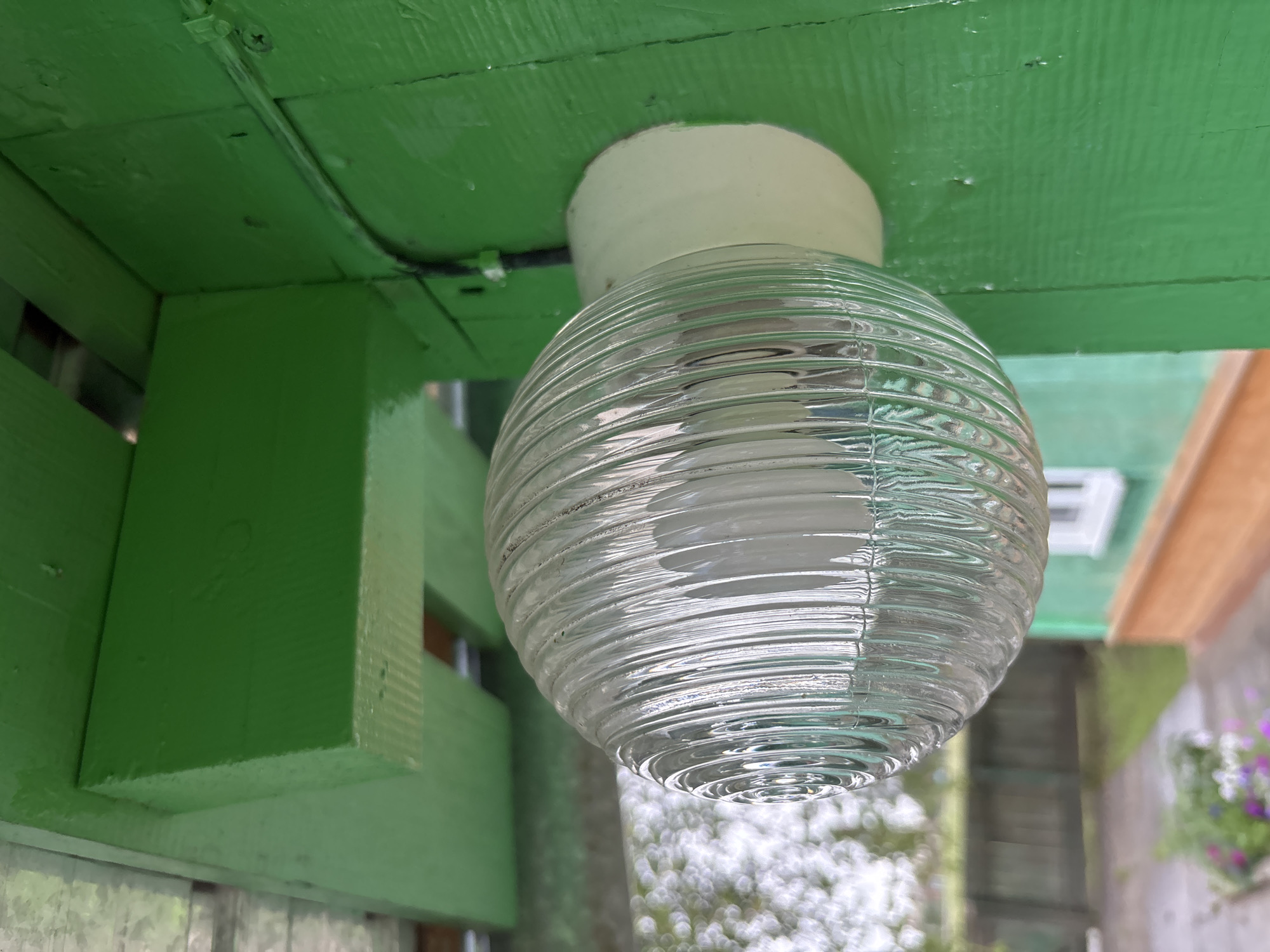 The image shows a close-up view of a light fixture mounted on a green wooden wall. The light fixture has a clear, ribbed glass cover with a white base. The glass cover is egg-shaped and appears to contain a bulb. The wooden wall is painted a vibrant green and shows some signs of wear, with visible grain and small imperfections. Above the light fixture, a green wooden beam is visible, also painted green. In the background, out of focus, is a blurry view of a yard with a wooden fence and some greenery. The overall impression is of an outdoor setting, possibly in a rural area. There is no indication of Gonja, Russia within the image itself.