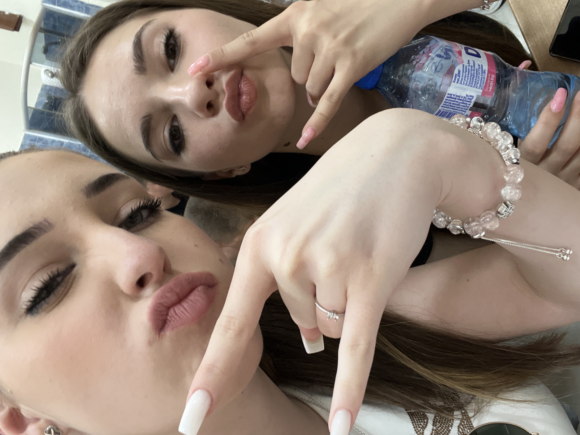 Two young women pose closely for a selfie. The woman in the foreground wears white nail polish, a ring on her ring finger, and a beaded bracelet. The other woman in the background holds a bottle of water with pink nail polish. They both purse their lips and use their hands to make a peace sign. The location is not discernible, but the bottle of water says "Vechernik 5L" which is Bulgarian for "Evening 5L". The water is bottled in Stara Zagora, Bulgaria.