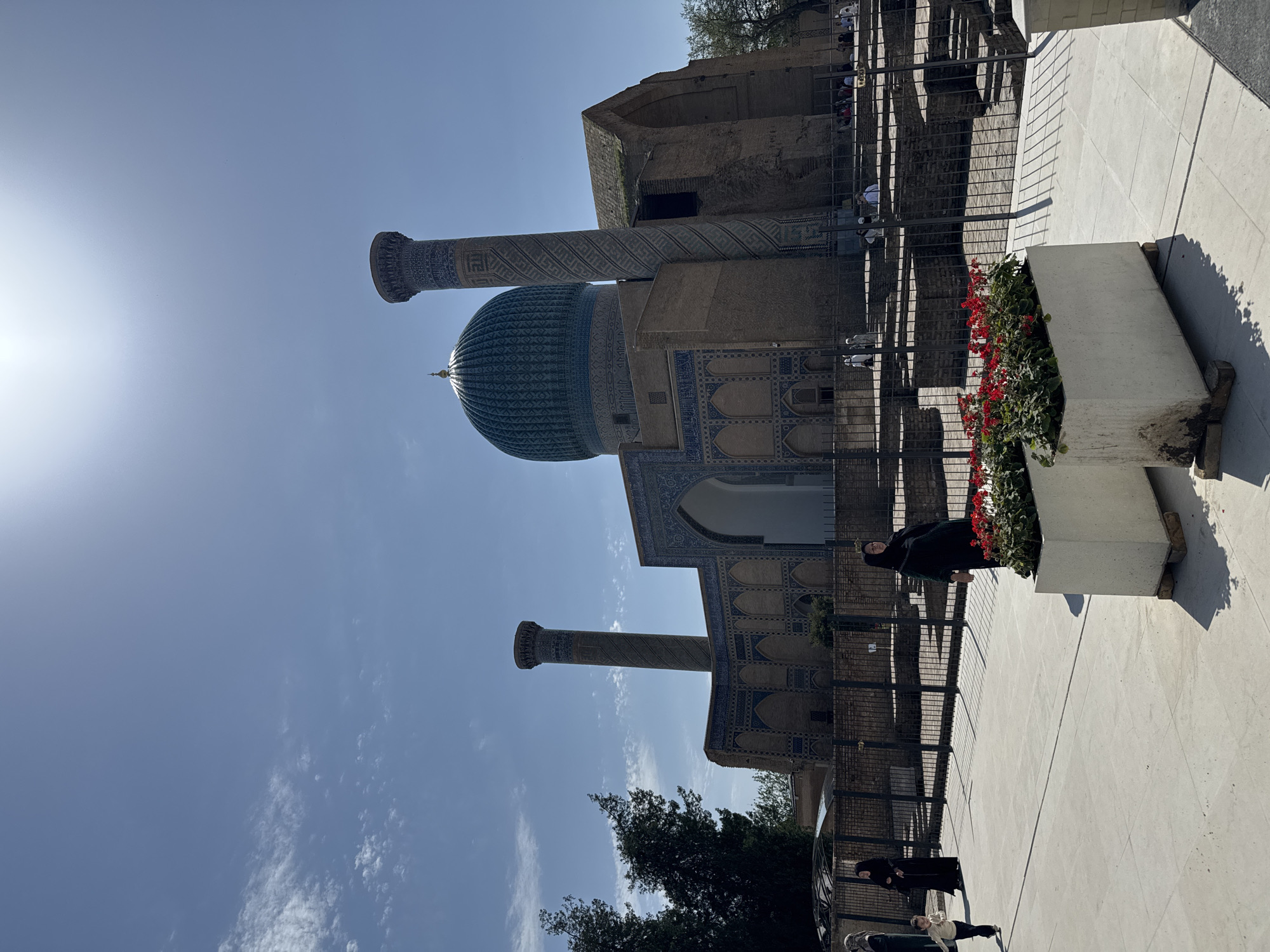 The image shows the Gur-e-Amir Mausoleum in Samarkand City, Uzbekistan. The mausoleum is a complex featuring a large dome and tall, cylindrical minarets. A woman in black stands near a large, white planter box filled with red flowers in the foreground. A black metal fence separates the viewer from the mausoleum. Other individuals can be seen in the background, near the mausoleum's structure. The sky is blue with scattered clouds.