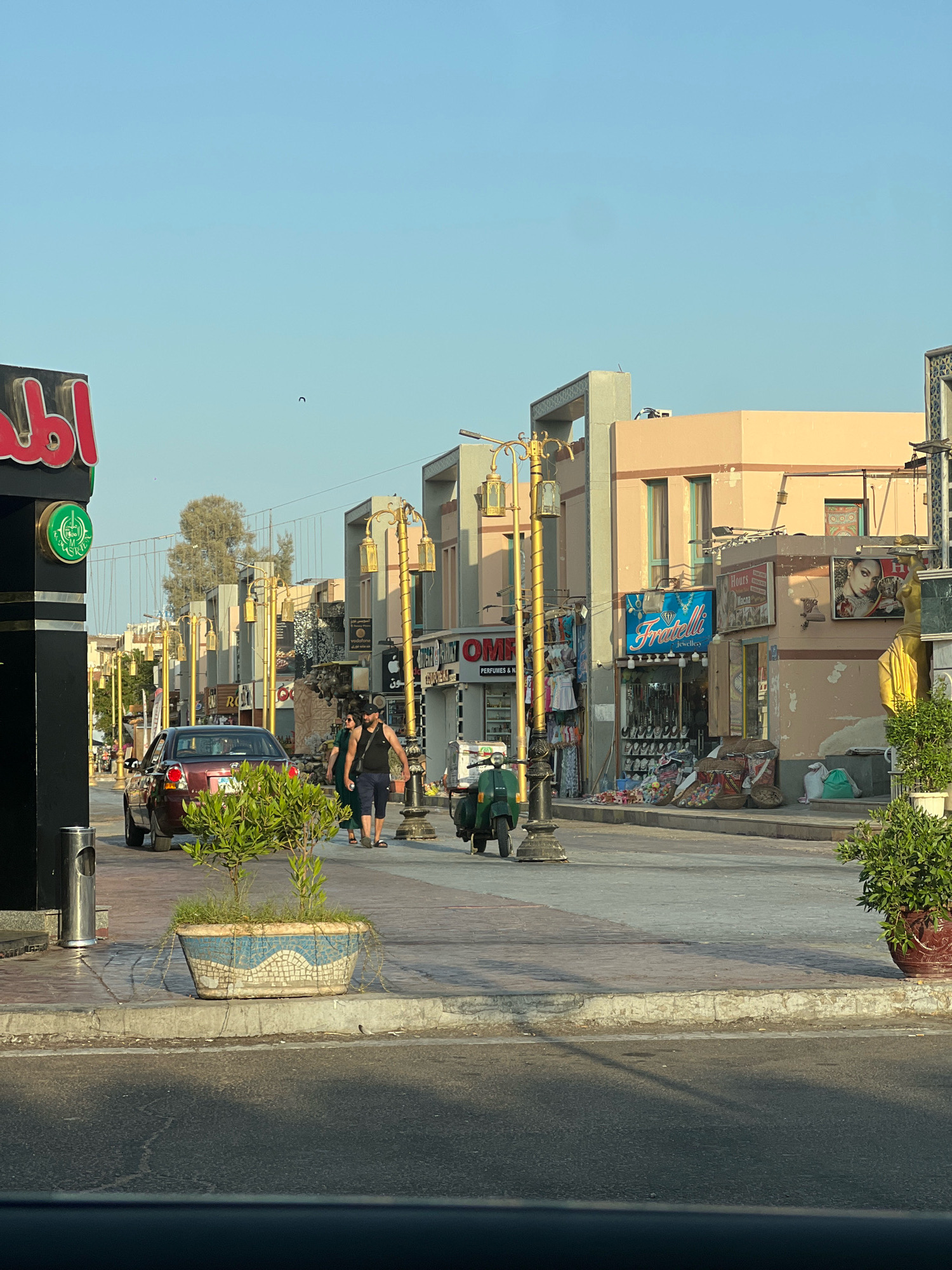 The image depicts a street scene in Sharm El Sheikh, Egypt, with buildings lining both sides of a paved road. The buildings are of various colors and architectural styles, featuring storefronts with awnings and signs. The sky is clear and blue.
On the left side of the street, a black sign with red Arabic lettering is visible, with a green circular logo below it. A red sedan is parked on the side of the road. A planter with a small green tree and decorative mosaic sides is positioned on the sidewalk in front of the car.
Several people are walking on the sidewalk. Two individuals, a man and a woman, are walking together near the center of the image. Further down the street, other pedestrians can be seen.
A green scooter is parked on the sidewalk near the center right of the image. Streetlights with ornate golden fixtures are positioned along the sidewalk. The storefronts display various goods, including clothing and jewelry. One shop sign reads "Fratelli Jewellery."
The overall scene suggests a commercial area with pedestrian and vehicle traffic. The lighting indicates it is daytime, likely late afternoon, given the shadows.