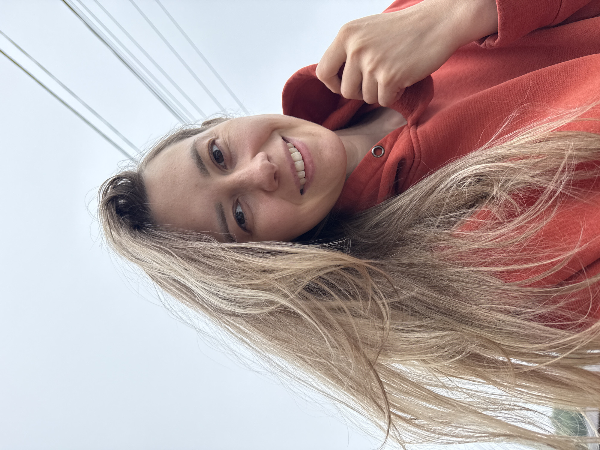 A young woman with long, light-colored hair and fair skin smiles broadly, looking upwards at the camera in a tilted selfie. She is wearing a reddish-orange hooded sweatshirt, with her right hand casually adjusting the collar near a silver grommet. Her dark eyes are open, and her smile reveals her teeth, conveying a cheerful emotion. The image is oriented such that her head is towards the bottom left and her hair flows across the frame.
The background consists of a bright, light gray sky, likely overcast, against which several thin, parallel lines are visible diagonally in the upper portion, suggesting power lines or bridge cables. This outdoor scene is set during the daytime in Chaikovsky, Russia. No visible text is present in the media file.