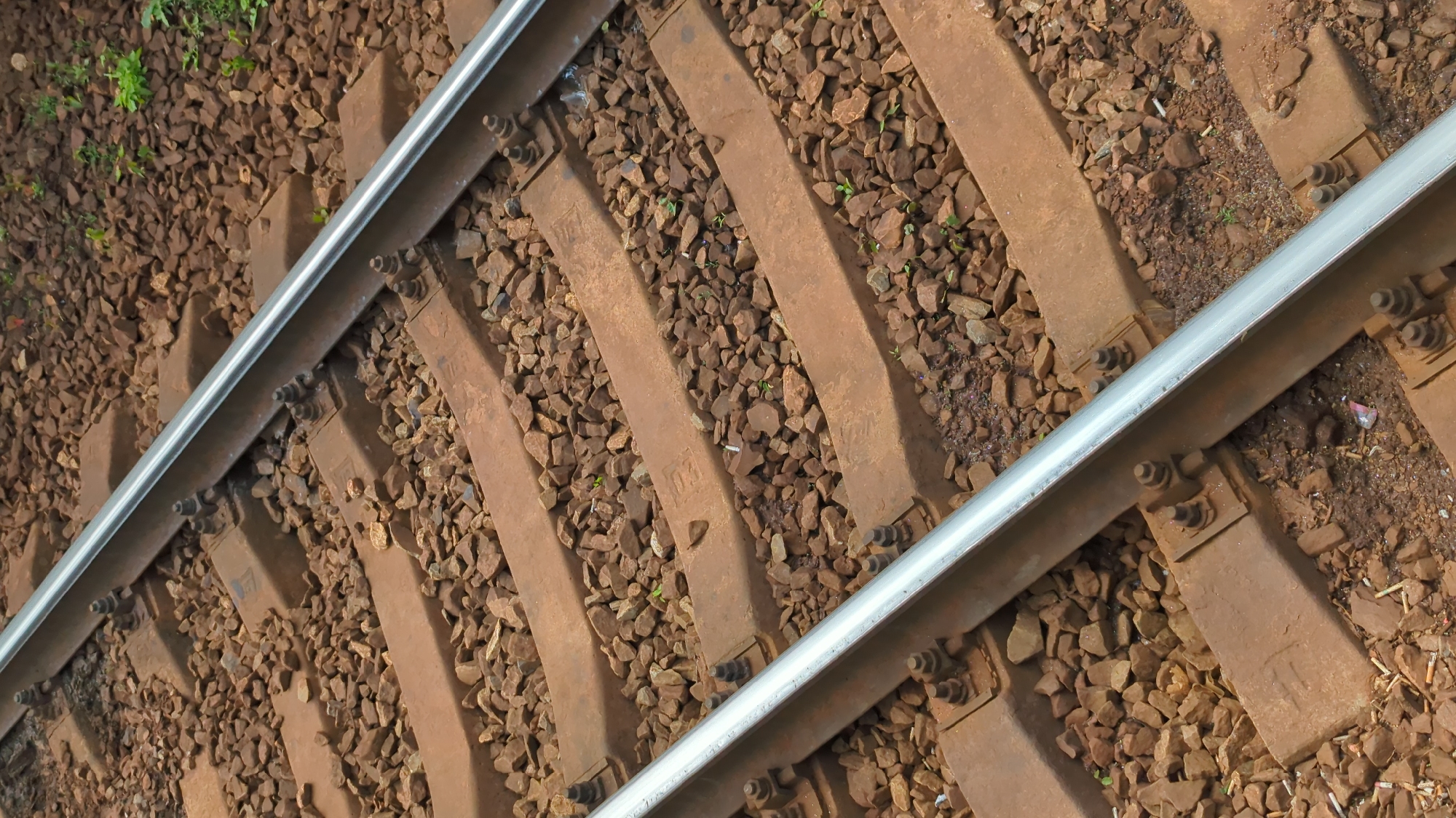 The image shows a close-up, high-angle view of a railroad track. Two parallel steel rails are visible, supported by numerous concrete railway sleepers, which are embedded in a bed of crushed stone ballast. The sleepers are secured to the rails by metal clips and bolts. Small green shoots are growing between the ballast stones. The scene is an outdoor environment, consistent with a railway line in Moscow, Russia, though no specific landmarks are identifiable. No people or animals are present in the image.