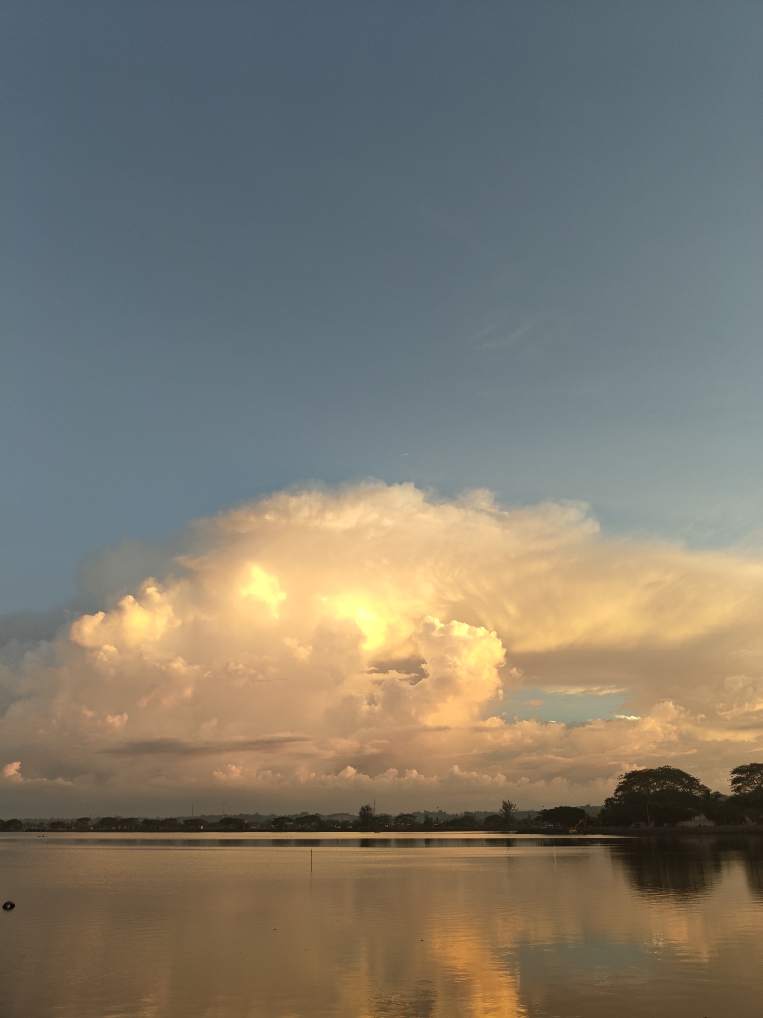 This is a landscape photograph of a large body of water reflecting the sky and a dramatic cloud formation at sunset. The sky is a gradient of soft blue at the top, transitioning to warm hues of yellow and orange near the horizon, illuminated by the setting sun. A large, voluminous cloud dominates the central portion of the sky, brightly lit from within by the sun, creating a luminous, almost golden appearance. The water in the foreground is calm and glassy, mirroring the colors of the sky and clouds. The horizon line is low, with a distant treeline and some structures visible on the land. This scene is characteristic of the waterside areas in Lhokseumawe, Indonesia, where expansive skies and bodies of water are common, especially during twilight hours, creating picturesque views.