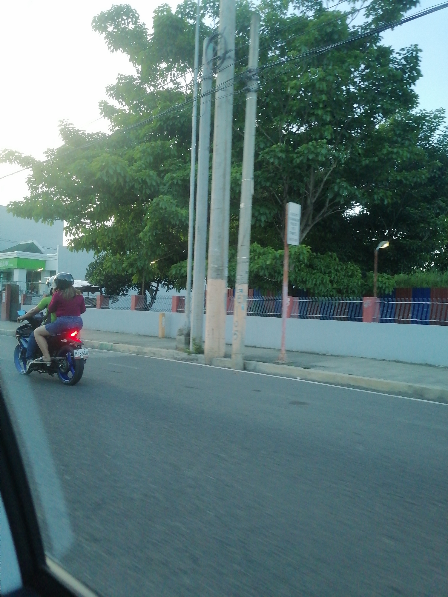 This image captures a street scene in Davao City, Philippines, likely near a BDO Unibank branch, during the late afternoon or early evening. The photograph is taken from a moving vehicle, evidenced by the slightly blurred foreground and the perspective from within a car on the lower left.
In the mid-ground, a dark blue motorcycle with blue wheel rims travels away from the camera on an asphalt road. Two individuals are on the motorcycle. The driver, wearing a green shirt and a black helmet, is partially obscured. The passenger, a female, is seated behind the driver, wearing a dark red long-sleeved shirt, blue denim shorts, and a black helmet. Her bare legs are visible, and she is wearing sandals. The motorcycle's red taillight is illuminated, and its Philippine license plate, "M486AW," is visible.
To the immediate right of the road, a sidewalk features several concrete utility poles with overhead power lines. Further down, a white, unreadable sign on a thin reddish-brown pole stands in front of a low white concrete wall topped by a fence composed of alternating blue and reddish-brown vertical slats. Dense green foliage and large trees are visible behind this fence, along with a street light pole whose lamp is illuminated.
In the left background, a light blue-grey building with a prominent green-branded entrance is visible. The partial word "BANK" on the green signage strongly suggests a BDO Unibank branch. Above and behind the structures, a bright, hazy sky is visible, with numerous large, green-leafed trees dominating the upper portion of the frame. The road itself is paved with asphalt, and a white solid line marks its edge on the right.
