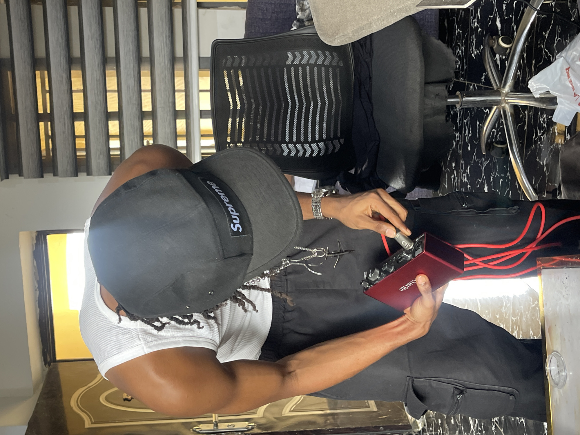 The image shows a person in black cargo pants and a white ribbed tank top, wearing a black baseball cap with a "Supreme" logo. The person is bending over and holding a small red audio interface, connecting a silver cable to it. Red cables are also visible. The person is wearing a silver watch and a silver necklace with a cross pendant. The background features a partially open door with intricate carvings, a black mesh office chair, and a window with horizontal blinds. The floor has a black and white marble pattern. The scene is likely indoors, possibly in a residential or office space within Abuja, Nigeria.