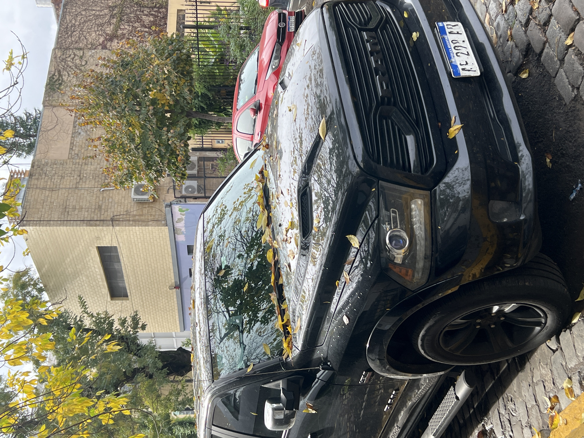 A black Dodge Ram pickup truck is parked on a cobblestone street in Buenos Aires, Argentina. The truck's windshield and hood are covered in fallen yellow leaves. A red Fiat is parked behind the truck. The license plate on the black truck reads "AE 299 FR". The background includes buildings, trees with yellowing leaves, and wrought-iron fencing. The weather appears overcast.