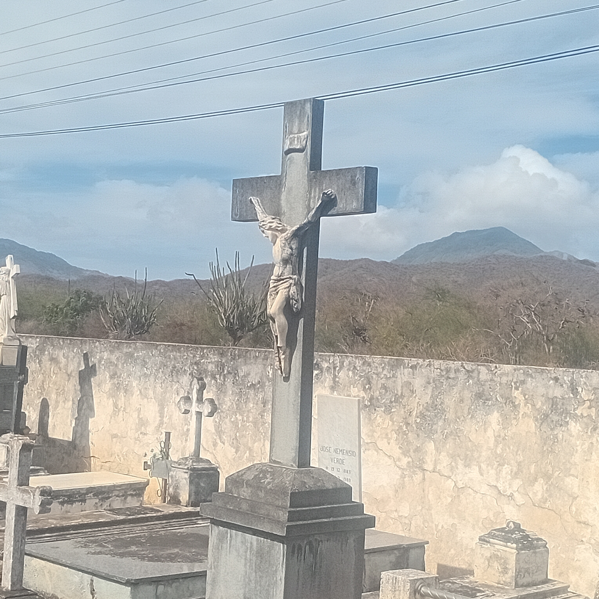 The image shows a concrete crucifixion statue in a cemetery, likely located in the city of Unknown, Unknown. The statue is weathered and features a figure of Jesus Christ on a cross. Behind the statue, there is a cracked concrete wall, and beyond that, a landscape of dry vegetation and hills with a mountain visible in the background. Several other tombstones and crosses are visible in the foreground. Overhead, power lines cut across the sky.