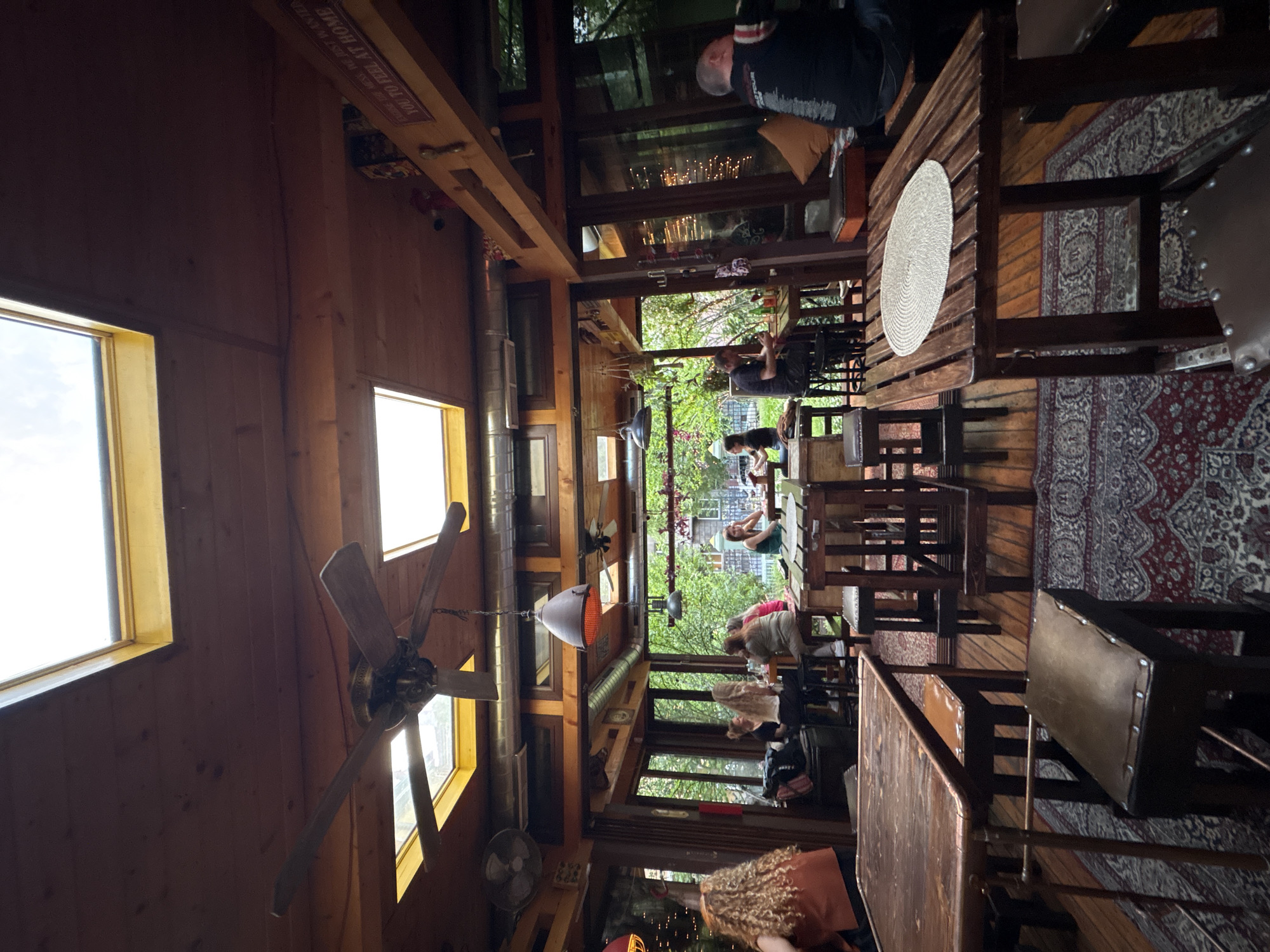 This image depicts the warm and rustic interior of a restaurant or cafe in Sofia, Bulgaria, illuminated by natural daylight. The setting is characterized by extensive use of wood, including paneled walls, exposed wooden beams, and dark wooden tables and chairs.
Numerous people are present, mostly seated at tables, engaged in dining or socializing. In the immediate foreground, the back of a person with curly hair wearing an orange top is visible. Further into the room, a man in a dark top and vest sits at a table, while other patrons, including a woman in a green top, are scattered throughout the space. Through large windows that line one side of the room, lush green foliage and glimpses of outdoor elements suggest an adjacent garden or an al fresco dining area.
Notable objects within the space include several vintage-style wooden ceiling fans, industrial-look light fixtures or heaters suspended from the ceiling, and exposed metal ductwork. The floor is adorned with patterned rugs in red and dark blue hues, adding to the cozy atmosphere. One wooden table features a white, circular woven placemat.
Visible text on a prominent wooden beam near the ceiling reads: "WELCOME TO THE MEN'S HUT. WE WANTEN YOU TO FEEL AT HOME." The time of day appears to be daytime, given the bright natural light filtering through the windows, and the overall mood suggests a relaxed and welcoming environment.