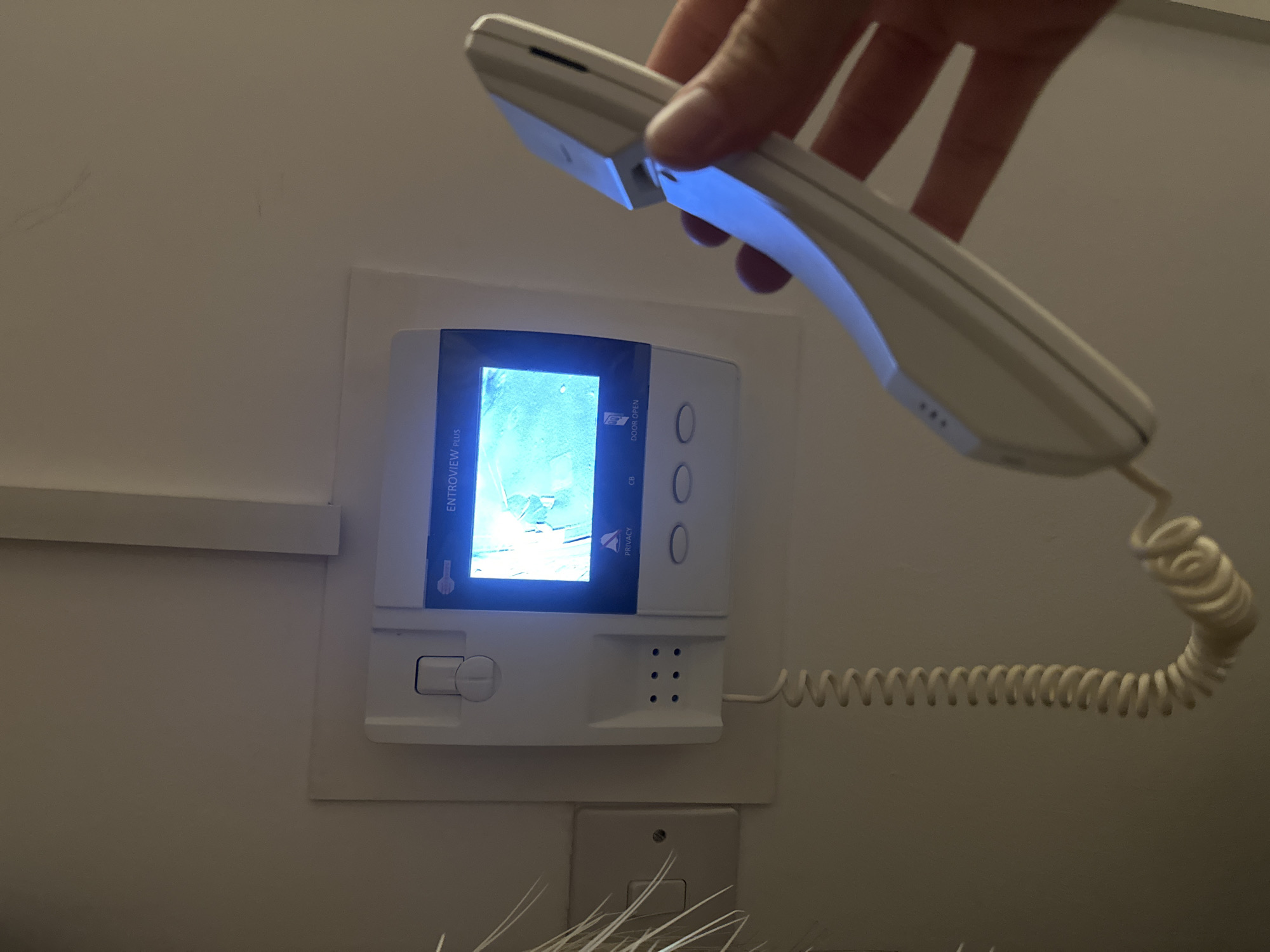 A hand holds a white handset receiver towards a white intercom system mounted on a light-colored wall. The intercom has a screen displaying a blurry image, several buttons, and a volume control. A coiled cord connects the handset to the intercom base. Below the intercom, a white electrical outlet is visible. The location is unknown.