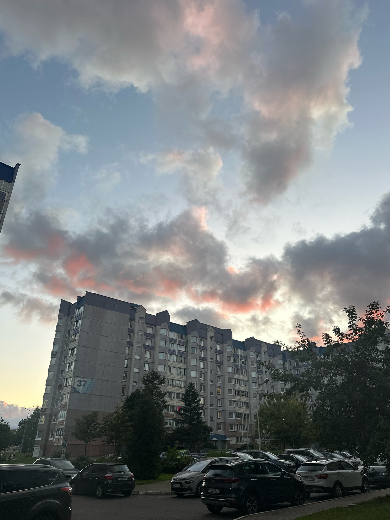 The image captures a residential building in Minsk, Belarus, bathed in the soft light of dusk. The sky is filled with dramatic clouds tinged with pink and orange hues, suggesting the setting sun. The building, likely an apartment complex, is tall and features multiple stories with numerous windows. A blue sign with the number "37" is visible on its facade. In front of the building, a street is lined with parked cars, mostly SUVs and sedans. Trees and other vegetation are scattered around, some of which partially obscure the building and the parked vehicles. The scene appears calm and ordinary, depicting a typical urban evening in Belarus.