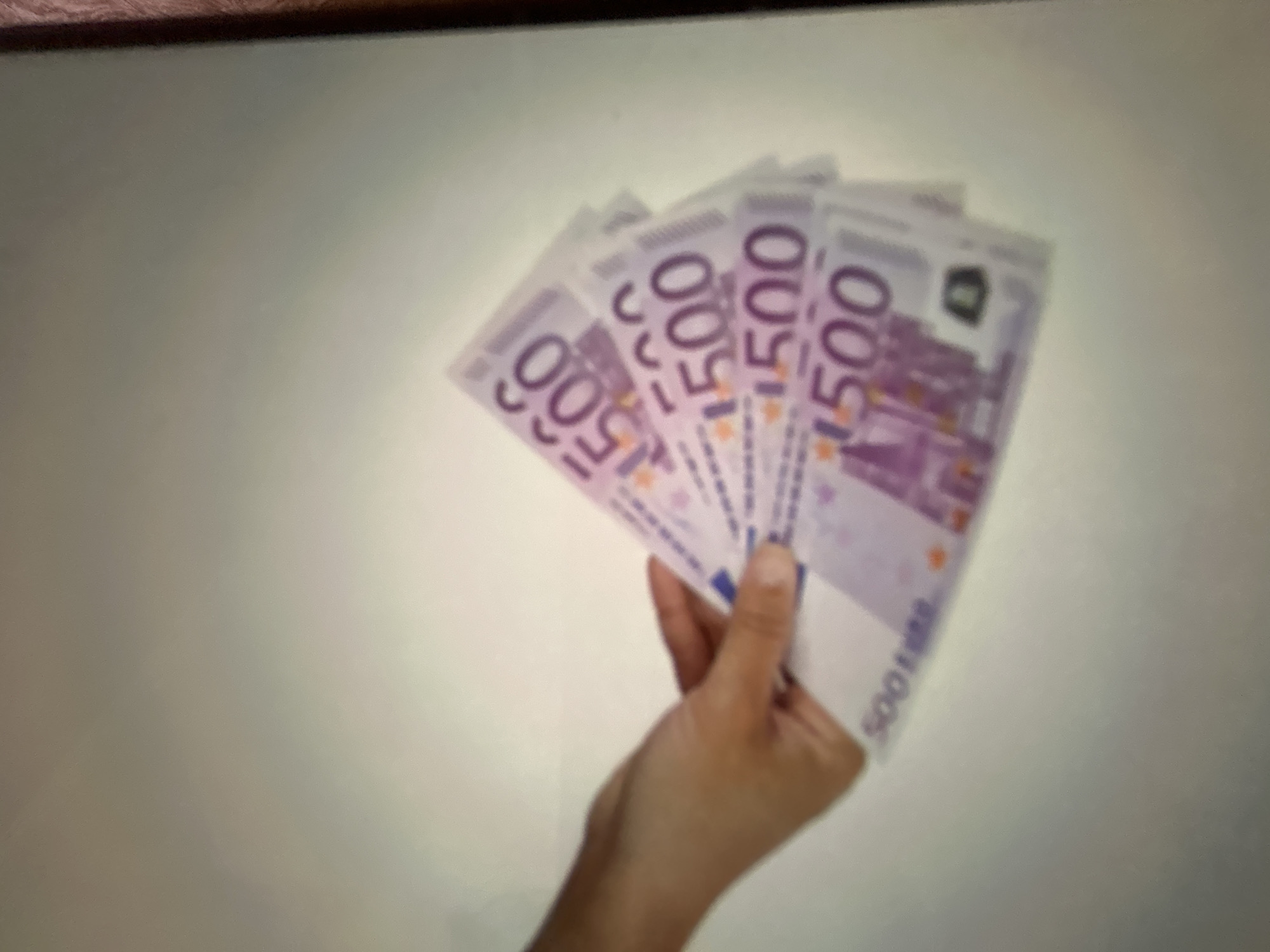A close-up, slightly blurred shot shows a person's hand holding a fanned-out stack of five 500 Euro banknotes. The notes are predominantly purple and feature the number "500" prominently displayed. The background is a plain, off-white surface. The scene is devoid of any recognizable Gelendzhik, Russia landmarks.