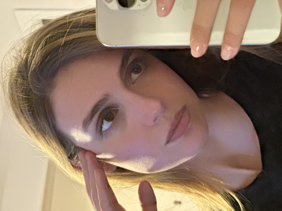 A close-up, self-portrait depicts a light-skinned individual, appearing to be a young adult female, indoors at an unknown location in the city of Unknown, Unknown. The individual possesses medium-brown hair with lighter highlights, brown eyes, and dark eyebrows. She is wearing a black, scoop-neck garment. Her right hand, exhibiting clear or light-pink nail polish, holds a light-colored smartphone (consistent with an iPhone Pro model) to capture the image; the phone's triple-lens camera array is visible on the right side. Her left hand is raised, with fingers touching the temple area of her head. Her gaze is directed generally towards the viewer. The background is blurred, indicating an indistinct indoor environment with artificial lighting creating shadows across her right cheek and neck.