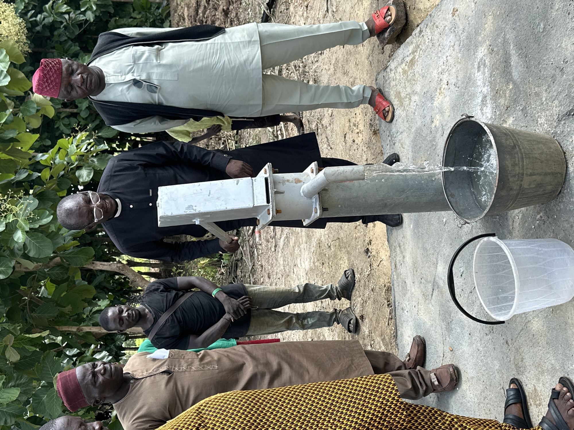 In Afogo, Nigeria, a group of men are gathered around a manual water pump. The pump, a silver metal cylinder with a lever arm and a spout, is actively dispensing water into a metal bucket. The ground is dusty earth, with patches of concrete where the pump is situated. Several men are dressed in traditional attire, including a man in a brown tunic and a red cap, and another in a light blue tunic and a dark vest, also wearing a red cap. A man in a black shirt and glasses appears to be operating the pump, while others observe. A clear plastic bucket also sits nearby. The background features lush green foliage, suggesting an outdoor, possibly rural or semi-rural, setting.