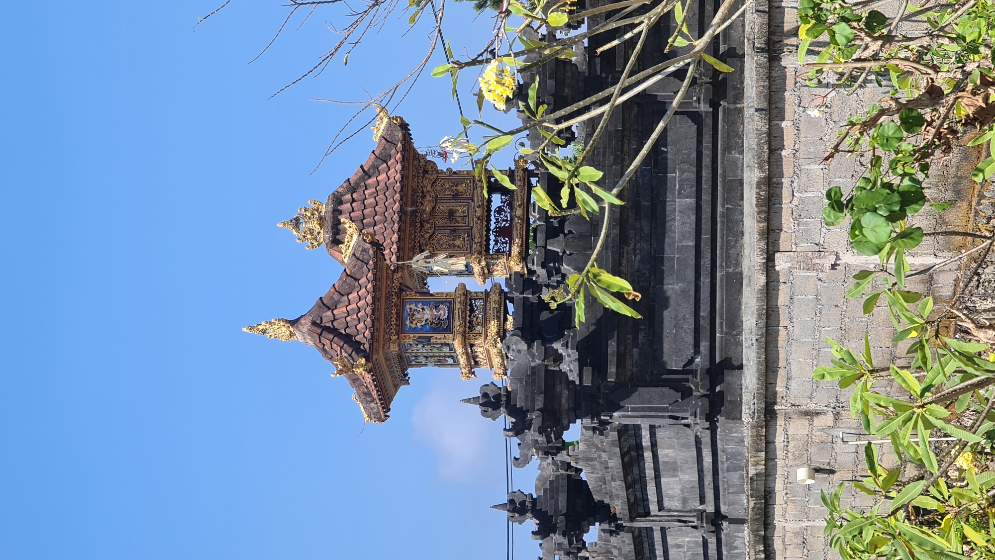 The image captures a view of traditional Balinese architecture against a clear blue sky. Two intricately decorated structures, likely part of a temple or shrine, stand atop a dark, stone wall. These structures feature ornate golden detailing and tiered, tiled roofs. Below them, the dark stone wall is adorned with decorative carvings and spires. In the foreground, lush green foliage and branches with a few yellow flowers partially obscure the view of the wall and structures. The lower portion of the image shows a lighter-colored brick wall. Based on visual cues and common architectural styles found in the region, this scene is likely located in Kuta Selatan, Indonesia, a popular tourist area known for its temples and traditional Balinese buildings. There are no people or visible actions occurring in the scene.