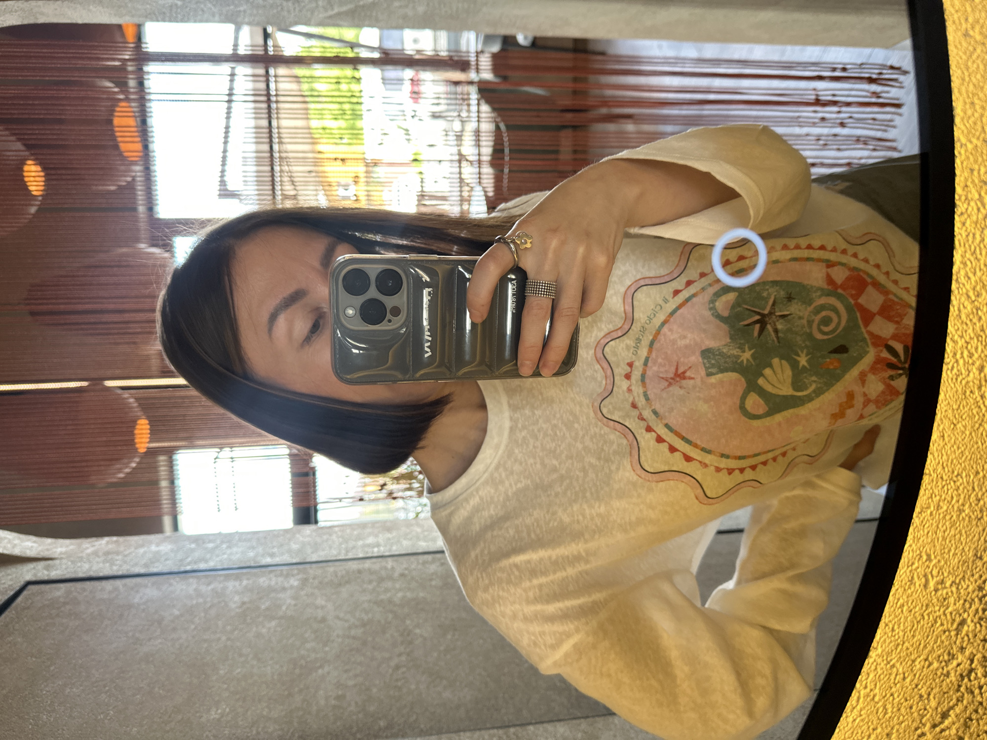 A female subject with dark brown bob-cut hair is depicted taking a mirror selfie in an indoor setting in Усады, Russia. Her reflection shows her holding an iPhone with a ribbed, translucent grey case displaying "VVV" and other text. She wears a light-colored, long-sleeved t-shirt featuring a circular graphic with a stylized pitcher, stars, and checkered patterns, and partially visible text "Il Cielo Picasso Il". Multiple rings are on her right hand: a multi-band silver-toned ring and a gold-toned ring with a small flower charm. The background, visible through the mirror, is a blurred interior space, likely a restaurant, characterized by vertical reddish-brown decorative strands, spherical orange light fixtures, and large windows revealing a bright outdoor scene with green foliage. The bottom edge of the mirror shows a textured gold-yellow surface.