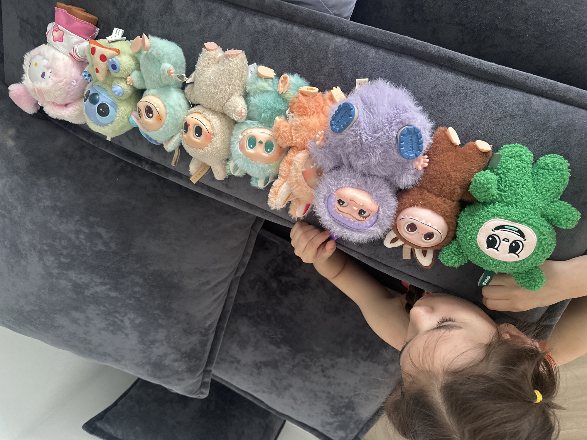 A young child lies on a grey sofa, with a line of plush toys arranged beside them. The toys are a variety of colors and designs, including a pink Hello Kitty, green Stitch, and several small monster-like figures with prominent eyes. The child is looking down, with their face close to the edge of the sofa. The scene appears to be indoors. There is no evidence to confirm the city of Kharnavy, Russia as the location.