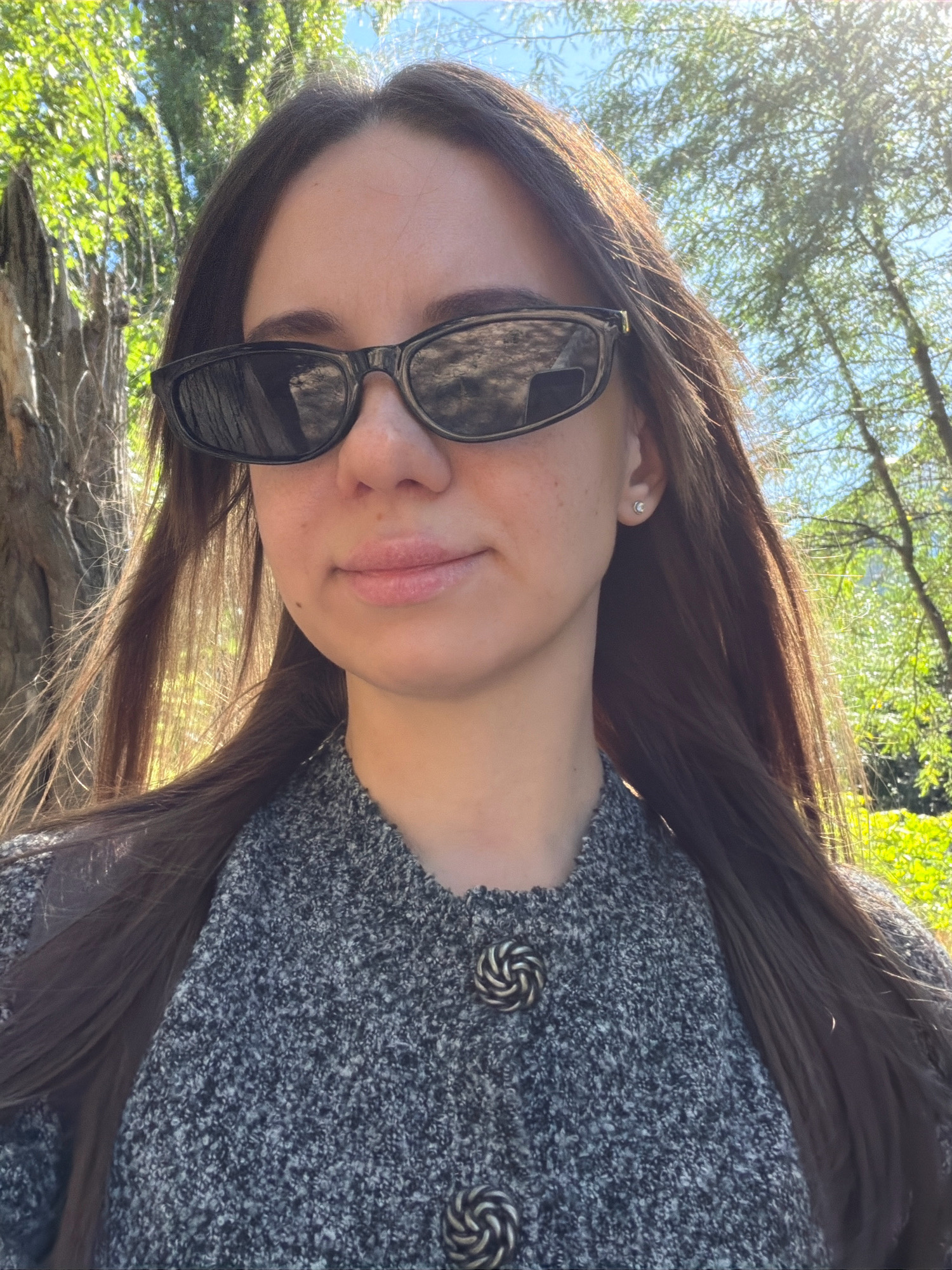 A medium shot selfie of a woman with long dark brown hair and fair skin is taken outdoors. She is wearing black sunglasses and a grey textured sweater with two decorative silver buttons. The background shows a bright blue sky and green trees, indicating a sunny day. The woman has a small stud earring in her visible ear. The lighting suggests it is daytime, with sunlight casting highlights on her hair and face. The overall impression is a casual outdoor portrait. The location is not discernible from the image itself, and no information is available to place it in Kyiv, Ukraine.