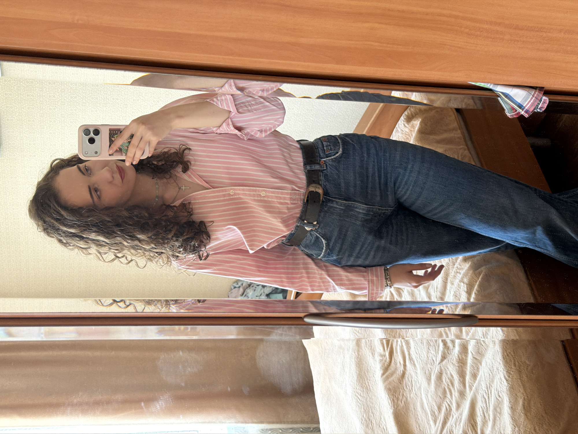 The image depicts a young woman taking a mirror selfie in what appears to be a bedroom. She is wearing a pink and white striped button-down shirt with cuffed sleeves and blue denim jeans with a black belt. She has curly brown hair and is wearing a silver cross necklace and a bracelet. In her right hand, she holds a pink phone case. The background shows a bed with beige bedding, a mirror with a wooden frame, and a light-colored wall with a subtle pattern. The overall scene suggests a casual, indoor environment. The location is the city of Zavodoukovsk, Russia.