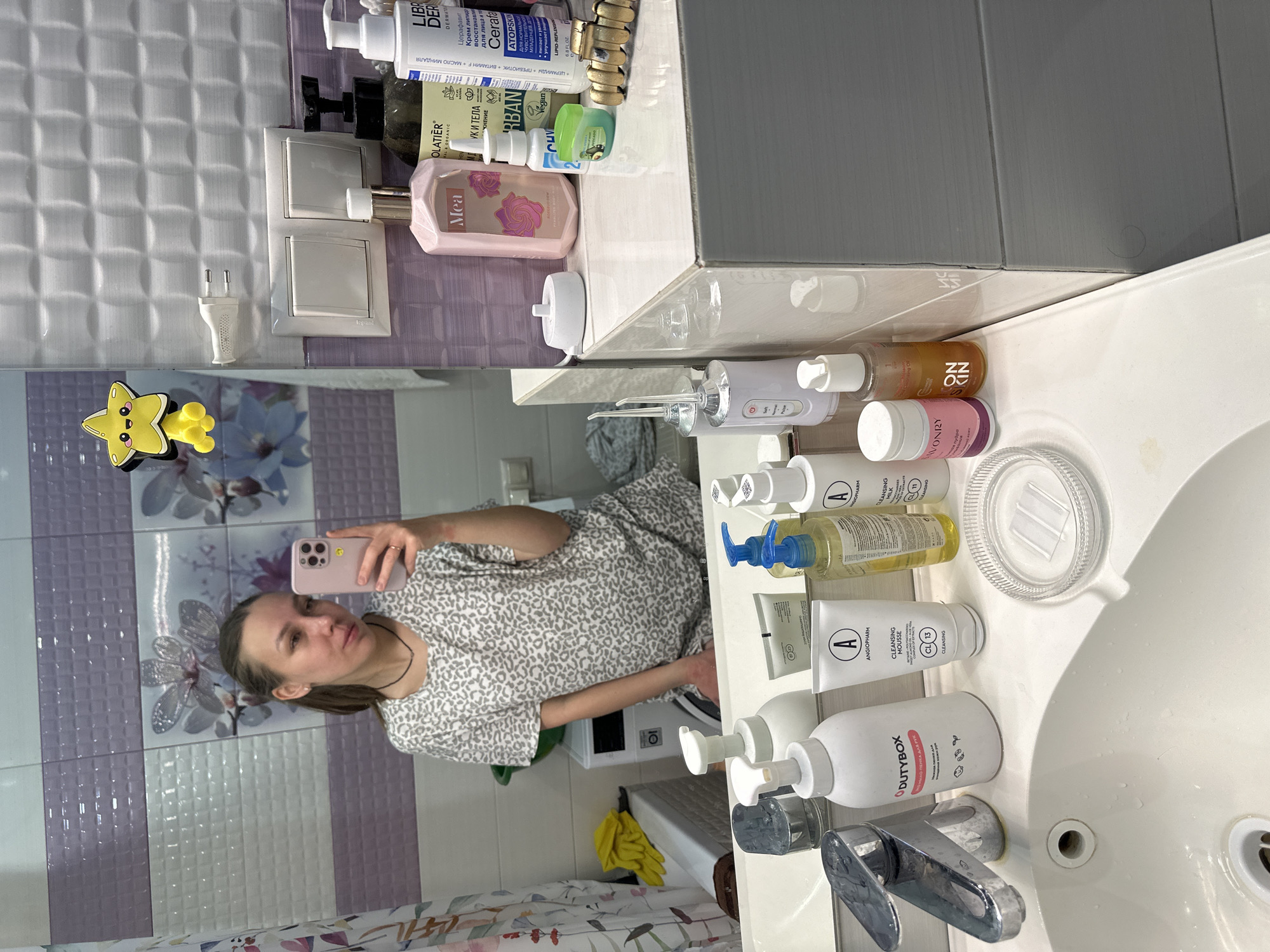 The image shows a selfie taken by a woman in a bathroom in Tomilino, Russia, reflected in a mirror. The photograph is oriented 90 degrees clockwise. The woman, with her brown hair pulled back, wears a light-colored, patterned top and is holding a light pink smartphone to take the picture.
The bathroom walls feature light and dark purple tiles, some with a textured pattern and others adorned with floral designs. On the counter by the sink, a collection of personal care products is neatly arranged. Visible items include a white Dutybox hand foam ("НО-ПЕНКА ДЛЯ РУК"), Angiopharm Cleansing Mousse ("CLEANSING MOUSSE CL13 CLEANSING"), and Angiopharm Cleansing Milk ("CLEANSING MILK CL11 CLEANSING"), along with a clear, round soap dish. Other products on the counter include an ON SKIN cleansing oil ("ON ZZ KIN"), a Savonry product ("SAVONRY"), and a white portable water flosser.
On the shelf above the sink, additional products are seen, such as Librederm Cerafavit Atopskin cream ("LIBR DER DERMATO Церафавит Крем липид восстанавл для лица и та Cerafa ATOPSKIN"), an Olatier Urban product ("OLATIER MAL & ORGANIC УК И ТЕЛА ЖНЕНИЕ URBAN Vegan"), and a pink "Mea" fragrance bottle. Two white electrical outlets are mounted on the tiled wall, and a yellow star-shaped clip is attached to the mirror. The sink itself is white ceramic with a chrome faucet. In the background reflection, a washing machine (likely LG branded) and a yellow cloth are discernible. The indoor lighting appears bright, possibly indicating daytime or well-lit evening conditions.