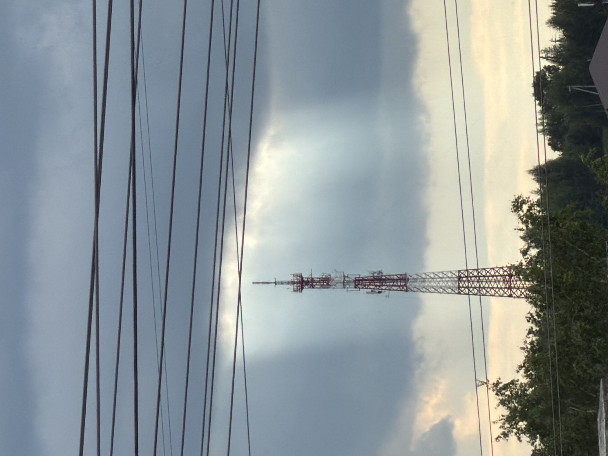 The image depicts a communications tower with red and white markings, viewed against a cloudy sky. In the foreground are several electrical or telecommunications wires, obscuring the left side of the view. The tower is located in or near a wooded area. Based on the location identified (Chaikovsky, Russia), it is likely a local telecommunications tower, but details of the specific environment or structures visible cannot be determined from the presented view.