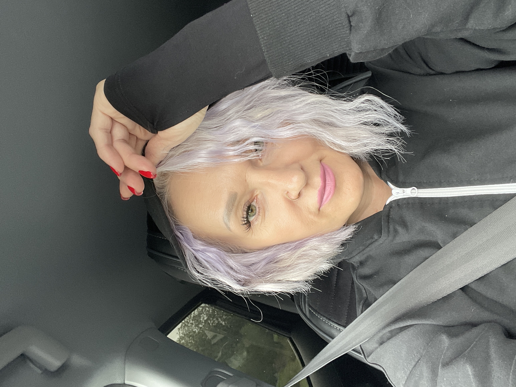 A close-up, forward-facing selfie captures a woman with short, wavy, lilac-tinged hair and light green eyes, wearing a black zip-up hoodie. She is seated in the driver's seat of a car, as indicated by the visible seatbelt. Her left hand is raised, touching the hood of her sweatshirt. The car's interior is dark gray, with a hint of the outdoors visible through the rear window, suggesting overcast weather. Her makeup includes pink lipstick and false eyelashes, and she is wearing a small hoop nose ring. The image is sharp, with good lighting and no discernible artistic manipulation. The exact location in Saint Petersburg, Russia, cannot be determined from the visual information provided.