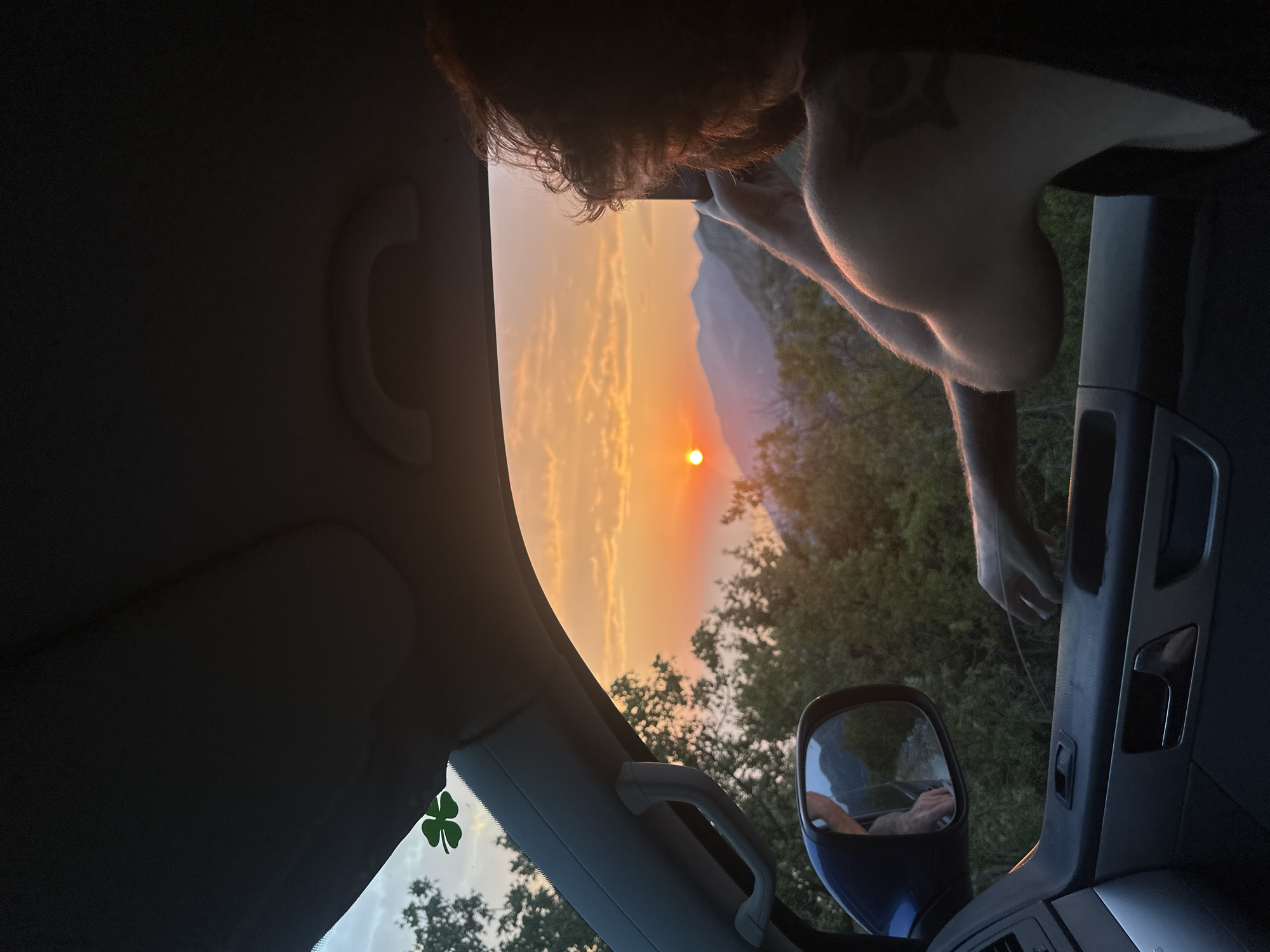 A person is seen from behind, leaning out of a car window in Gluhi Do, Montenegro, to photograph a sunset over mountains. The sky is filled with vibrant orange and yellow hues as the sun dips below the horizon. Lush green trees and rugged mountain peaks form the backdrop. Inside the car, a rearview mirror reflects the road and surrounding landscape. A small green clover sticker is affixed to the car's windshield. The person is wearing a black tank top and has a tattoo on their shoulder.