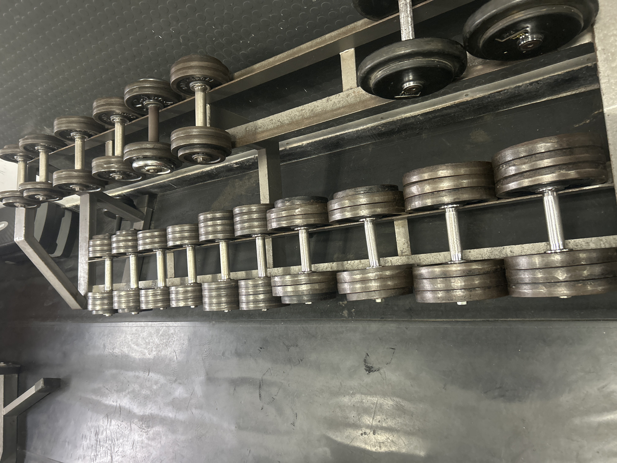 The image shows a rack of dumbbells in a gym setting. The dumbbells range in size, from smaller ones on the left to larger ones on the right. They are arranged on a metal rack with two tiers. The background includes a dark, textured wall and a gray floor with some smudges and markings. The lighting is somewhat dim, and the overall impression is of a functional, well-used workout area. No people are visible. There is no information about the city of Zlatograd, Bulgaria from the image.