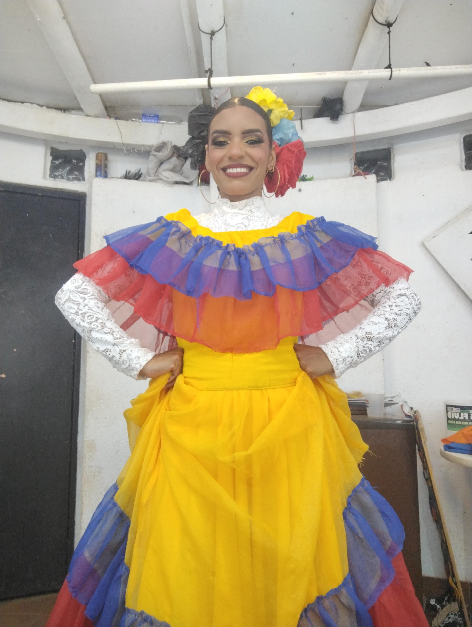 A woman is standing indoors wearing a traditional Venezuelan dress in the colors of the national flag: yellow, blue, and red. The dress features a layered, ruffled neckline and skirt. Her sleeves are white lace, and she has a floral headpiece in similar colors. She is smiling and has her hands on her hips.
The location appears to be a backstage or dressing room, possibly in Porlamar, Venezuela, based on the traditional Venezuelan attire. The background is cluttered with items such as a door, storage cabinet, and what appears to be various supplies and clothing items. The walls are white and show signs of wear and tear. There are exposed pipes and makeshift storage areas visible near the ceiling.