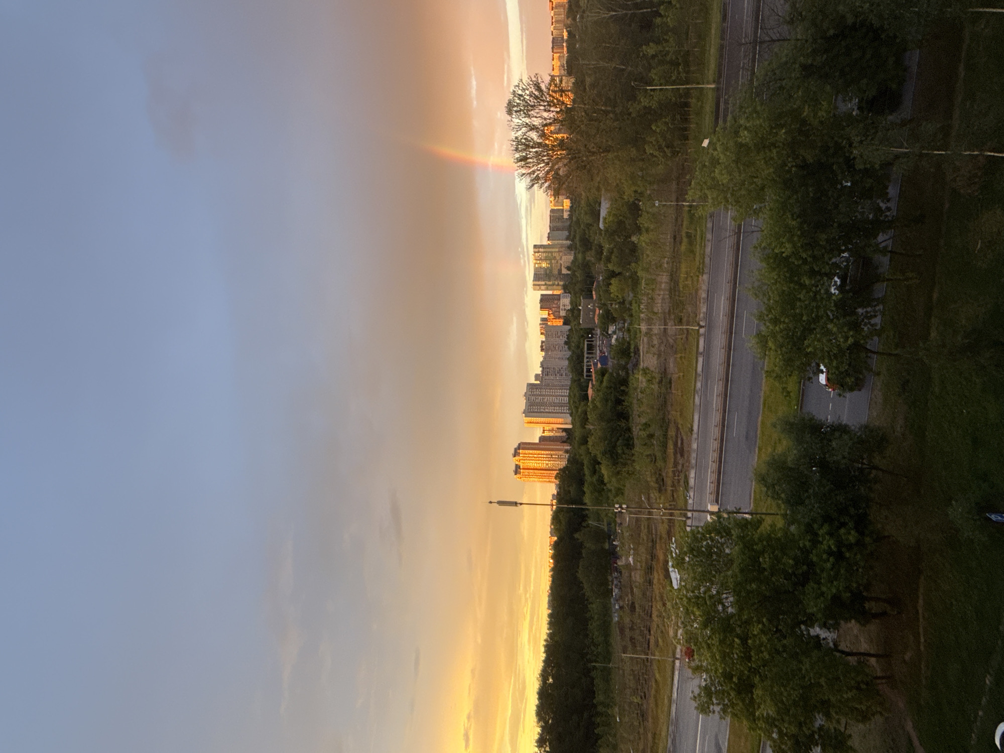 The image shows a wide view of a cityscape with a sunset. The sky is a gradient of light blue at the top, transitioning to orange and yellow hues at the horizon, with a visible rainbow arc. In the middle ground, there is a dense line of green trees, behind which are several multi-story buildings, appearing to be apartment complexes, illuminated by the setting sun. A tall, slender tower with antennas stands in front of the buildings. A highway runs horizontally across the lower part of the image, with a few cars visible on it, some appearing to be white, red, and black. The foreground consists of more trees, some of which are dark green, and an area of grass or low vegetation. The scene appears to be an urban or suburban environment, likely on the outskirts of a city given the presence of both dense greenery and residential buildings. The light suggests it is late afternoon or early evening. While the image is from Moscow, Russia, specific landmarks are not identifiable from this vantage point. There are no people clearly visible.