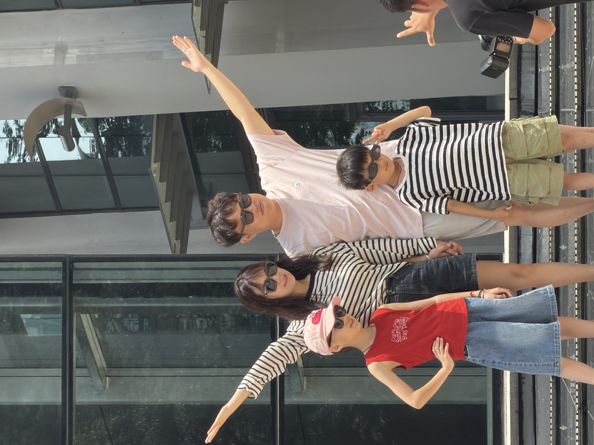 In the Yuzhong District of China, a family of four poses for a photograph in front of a modern building with large glass windows and a light gray facade. The family consists of two adults and two children, all wearing sunglasses. The adult male stands on the right, wearing a light pink t-shirt and raising his right arm. The adult female stands to his left, wearing a black and white striped long-sleeved shirt and raising her left arm. A young boy in the foreground, on the left side, wears a red tank top and denim shorts, with his left hand on his hip and his right arm raised. Next to him, a younger boy is dressed in a black and white striped t-shirt and khaki cargo shorts, making a peace sign with his right hand. In the background, a photographer, partially visible on the far right, is capturing the family's photo, holding a camera with a flash attachment. The scene is set on outdoor steps leading up to the building entrance.