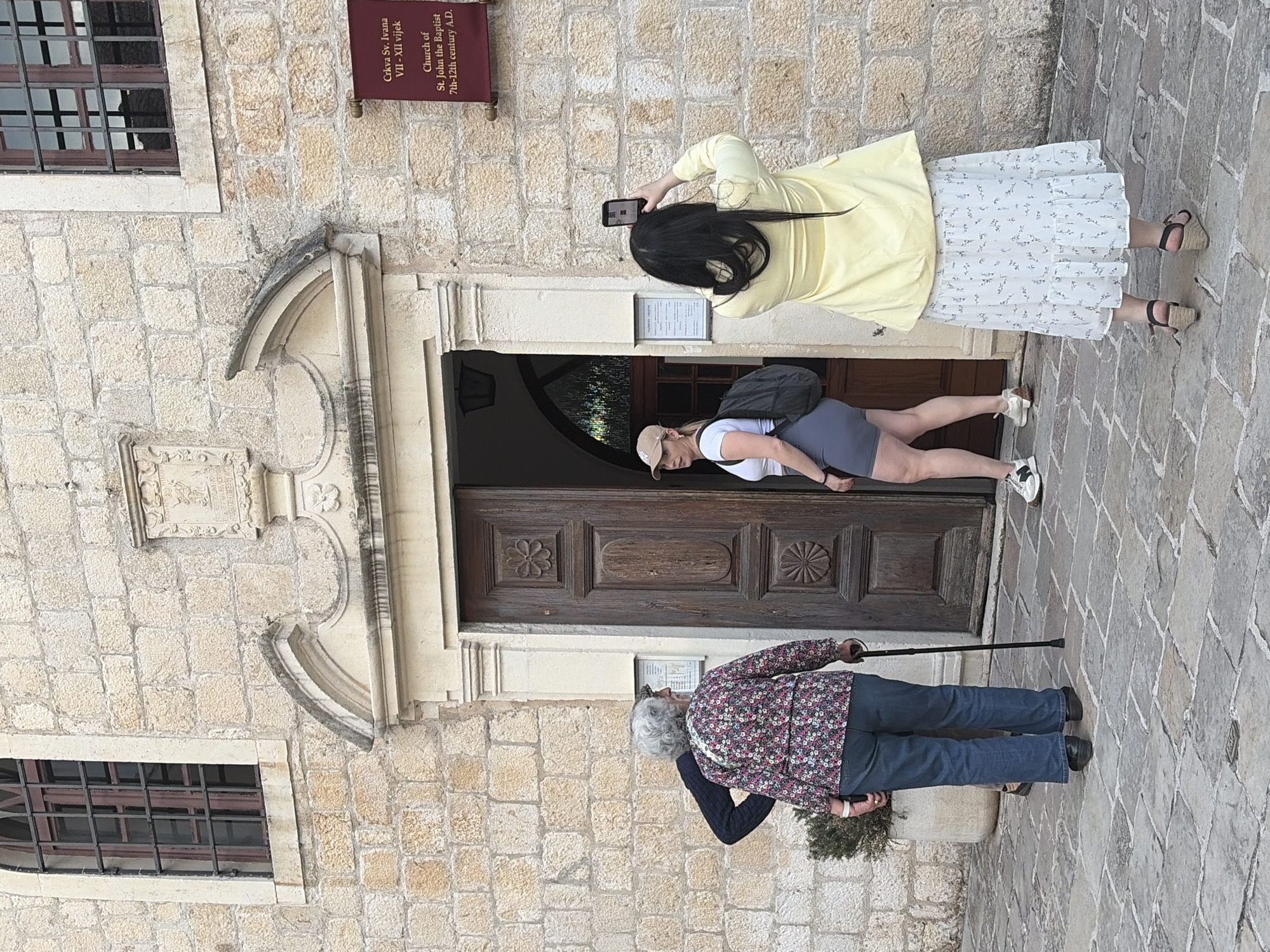 Here is a detailed description of the image:
**Scene:** The image shows the Church of St. John the Baptist in Budva, Montenegro. This church is a historic stone building.
**People:**
* An elderly woman in a floral patterned jacket, blue jeans, and black shoes is standing with a cane.
* A young woman in a yellow top and floral skirt is taking a picture with her phone.
* Another woman is wearing a white top, grey shorts, sneakers, and a backpack.
**Subjects:** The main subject is the church itself, marked by a sign indicating its history.
**Objects:**
* The church has an arched doorway and barred windows.
* The woman taking the photo is using a smartphone.
* The elderly woman is holding a cane.
* The woman wears a backpack.
**Actions:**
* One woman is taking a photo of the church.
* The elderly woman is standing and leaning on her cane.
**Interactions:** The women are all near the church, likely as tourists visiting the site.
**Environment:** The church is set within a stone-paved area. The scene is outdoors and appears to be during the day.