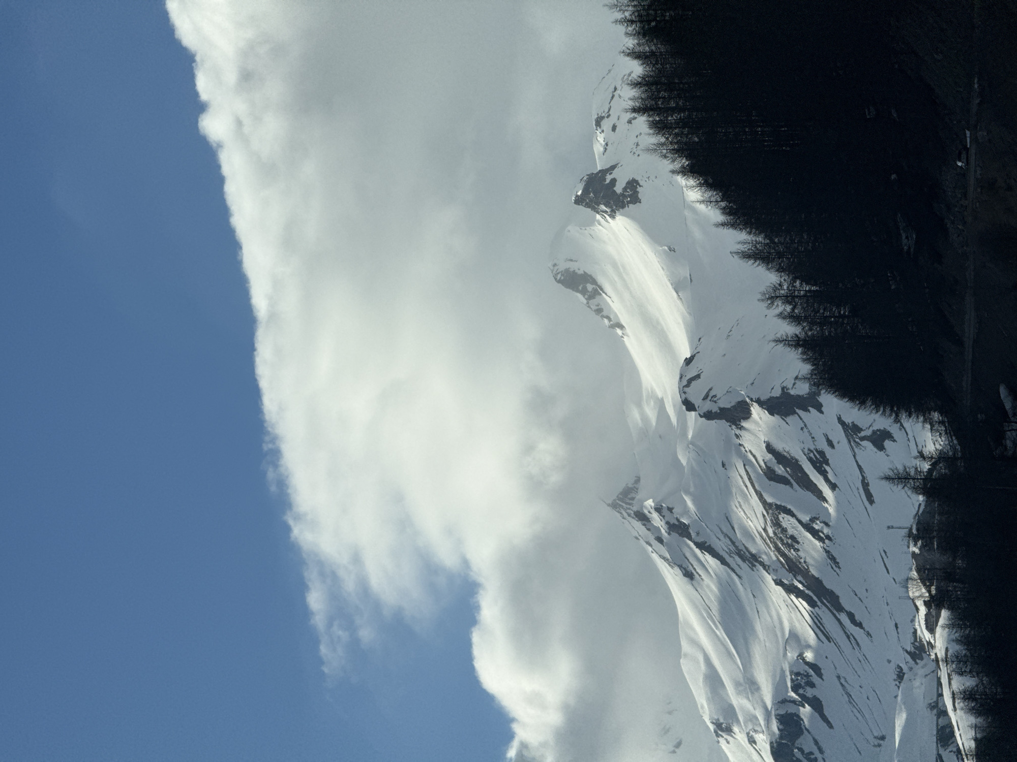 The image shows a view in Le Monêtier-les-Bains, France, featuring a snow-covered mountain range under a partially cloudy, blue sky. The mountainside is partly covered in dark evergreen trees along its upper reaches. Patches of snow are visible, reflecting sunlight. Clouds dominate the upper-central portion of the image, contrasted against the clear blue sky to the left.