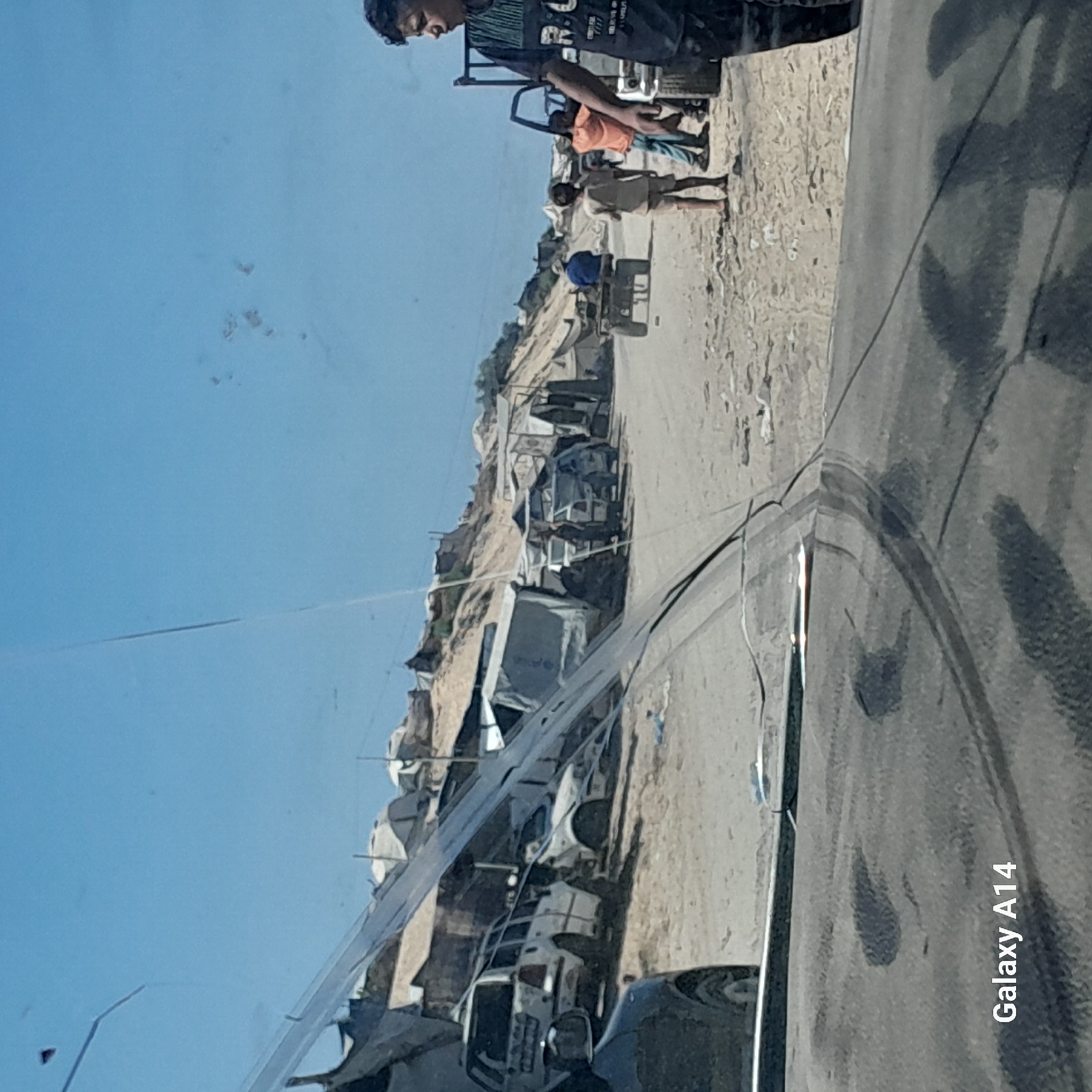 This image depicts a scene in Deir el-Balah, Palestinian Territories, showing a dirt road lined with makeshift shelters and tents, suggesting a refugee camp or informal settlement. Several vehicles are present, including white cars, one with its rear end visible and appearing somewhat damaged or abandoned, and another white station wagon. In the background, a sandy hillside is visible with more tents and structures.
On the road, several individuals are visible. In the foreground, a young boy in a dark t-shirt with graphics is looking to the right. To his left, another child stands, facing away from the camera. Further down the road, a person in an orange shirt and blue pants is standing near a truck. Another individual, wearing a blue shirt and a hat, sits on a cart-like vehicle. A person in a light-colored shirt and shorts stands near this cart. The overall environment is dusty and appears to be a warm, sunny day. A large crack is visible on what appears to be a car windshield in the foreground, obscuring parts of the scene. The bottom left corner of the image displays the text "Galaxy A14".