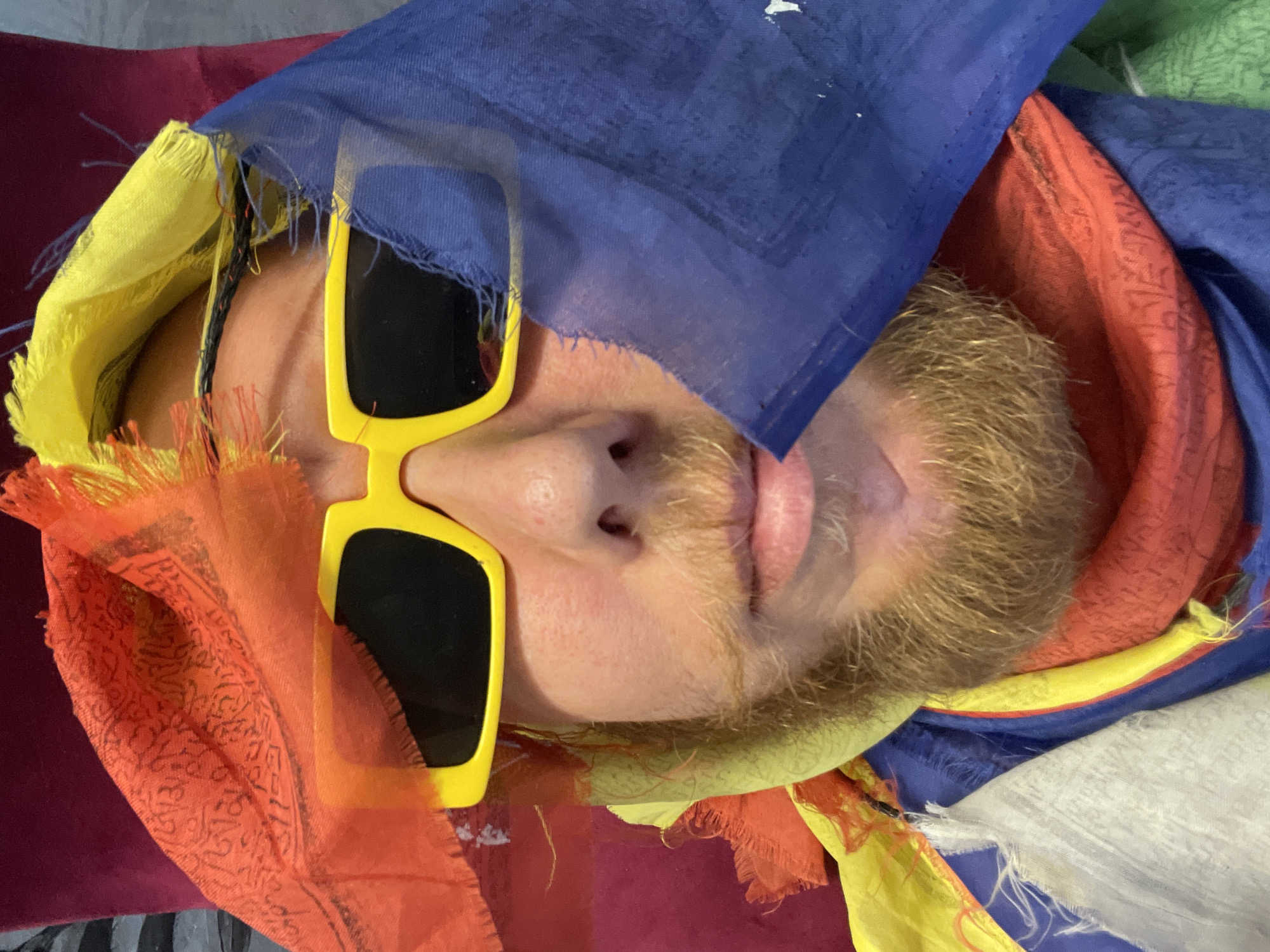 A person wearing bright yellow rectangular sunglasses and a full beard is covered in prayer flags. The flags are red, yellow, blue, and white, and they are torn and frayed. The person's face is partially obscured by the flags. The background is a dark red surface. The image is a close-up selfie. The location is not visible in the image.