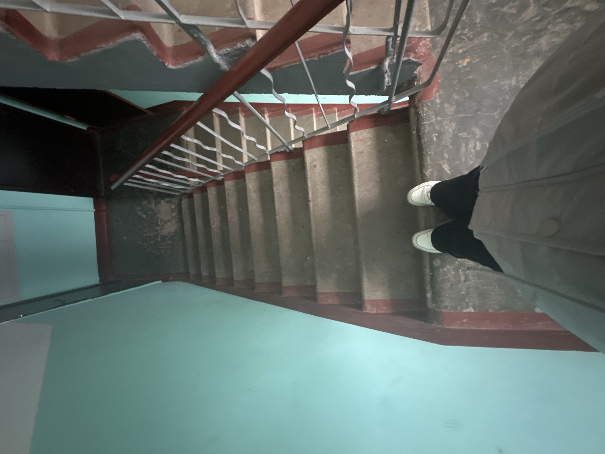 The image captures a top-down view of a person standing at the top of an indoor staircase, looking down. The setting appears to be an older, utilitarian building, consistent with post-Soviet architecture often found in locations like Chaikovsky, Russia.
In the foreground, the lower body of a person is visible, wearing black trousers, white sneakers, and a light grey jacket. The person is standing on a landing made of dark, worn concrete or a similar material.
The staircase itself is constructed from concrete, with individual steps featuring worn, dark red painted edges. A metal handrail runs down the right side of the stairs, comprising a dark red top bar and grey metal balusters with decorative, wavy elements. The walls lining the stairwell are painted in two distinct colors: a vibrant light blue or teal on the lower sections and a darker grey or black on the upper sections and ceiling.
The overall scene suggests a dimly lit interior, possibly relying on artificial light, as no windows are visible. There is no discernible activity beyond the person standing on the landing. The building materials and paint condition indicate age and wear. No text is visible in the image.