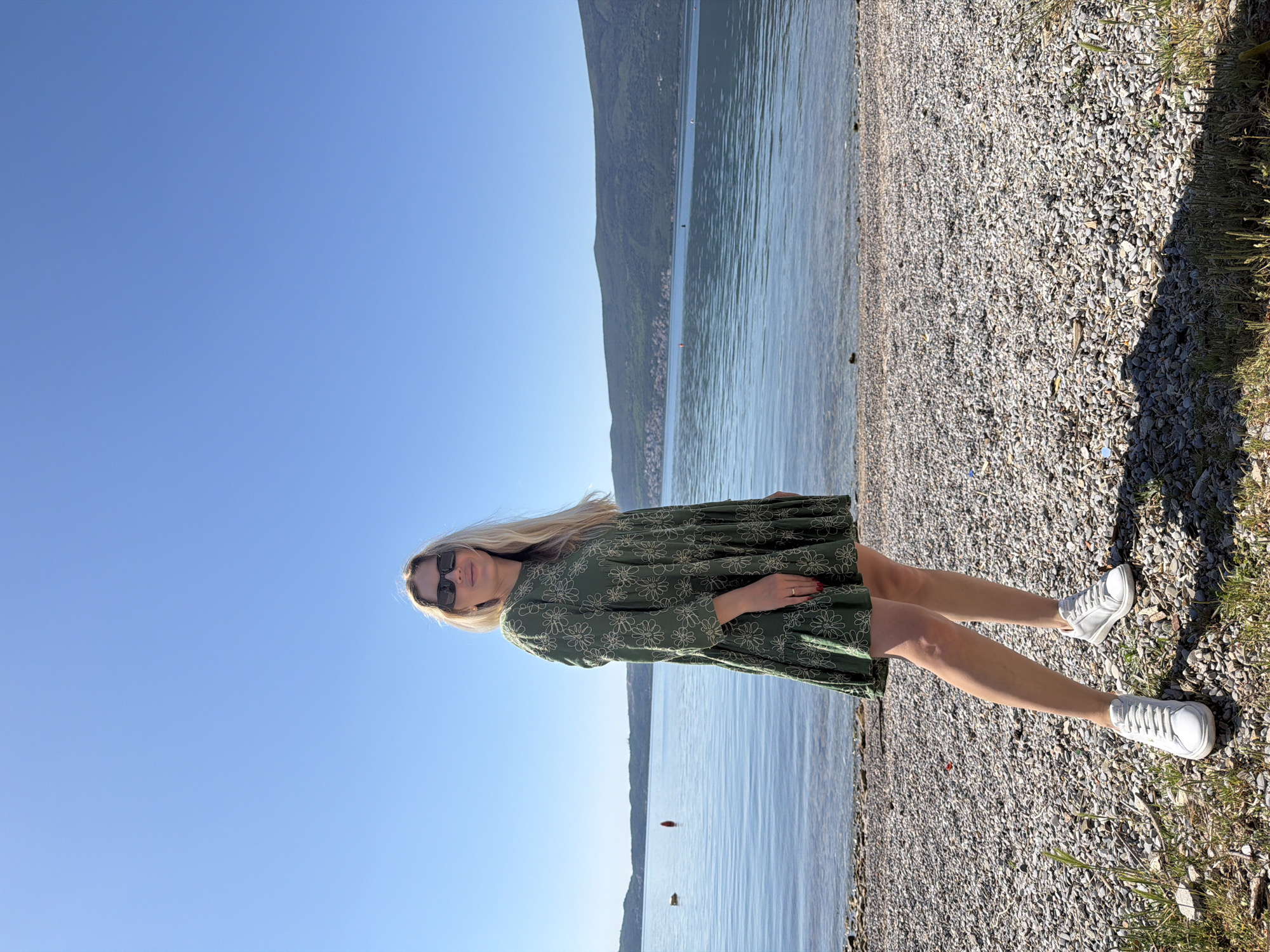 A woman stands on a pebbly beach beside a body of water. She has blonde hair, is wearing sunglasses, and a green floral dress. She is also wearing white sneakers. In the background is a mountain range and a clear blue sky. The location is Bijela, Montenegro.