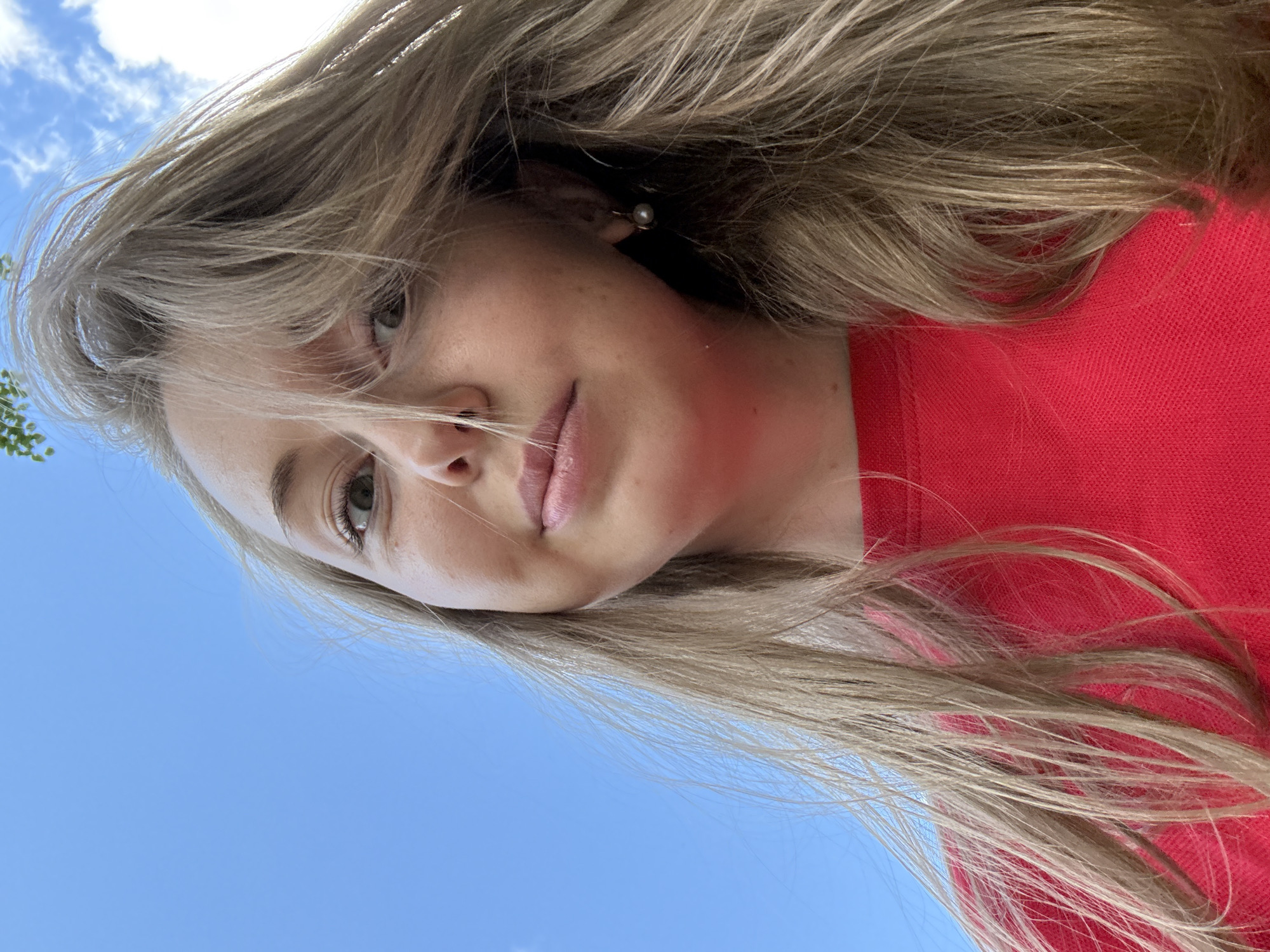 A woman with long, blonde hair is wearing a red shirt. She is outside, as evidenced by the blue sky in the background. The location is not specified.