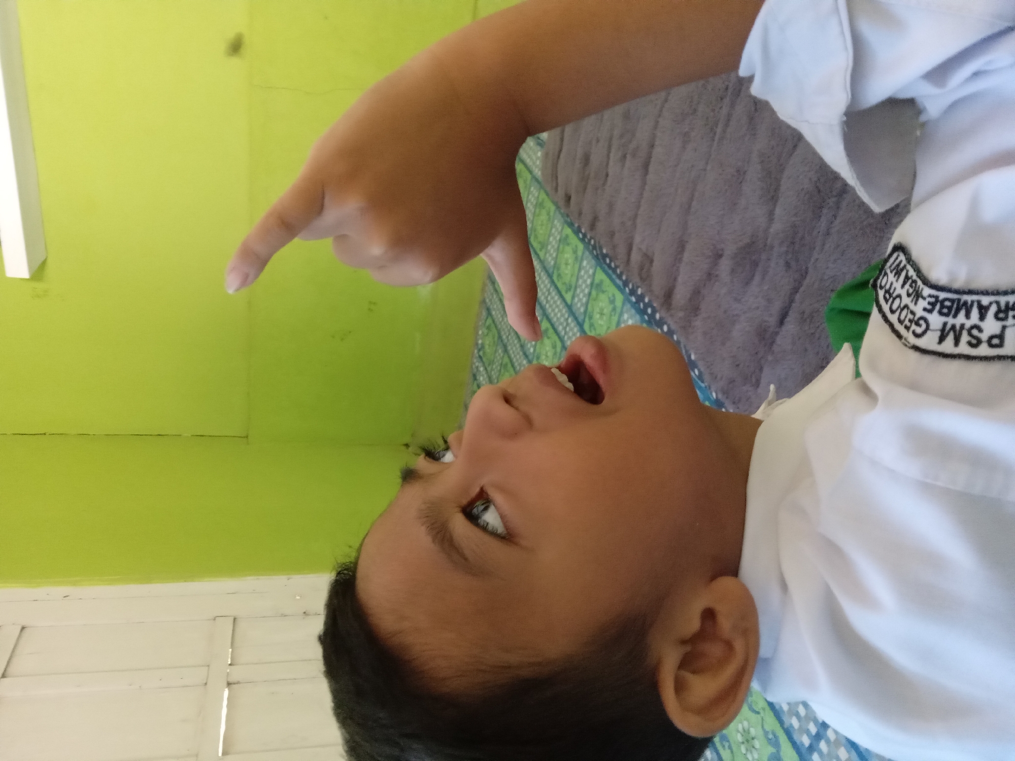 A young boy, likely Indonesian, wearing a white school uniform with a badge that reads "PSM GEDORO" and "BRAMBE NGAWI", looks up and to the right with his mouth open. He has dark hair and brown eyes. His right arm is raised, and his index finger and thumb are extended in a gesture resembling a shaka or "hang loose" sign. The boy is indoors, in what appears to be a room with a light green wall. A white wooden door is visible on the left side of the image, and a white fluorescent light fixture is mounted on the wall near the ceiling. A grey blanket or rug is spread on the floor, with a patterned mat beneath it. The overall scene is casual and appears to be taken in a residential or school setting in Kedunggudel, Indonesia.