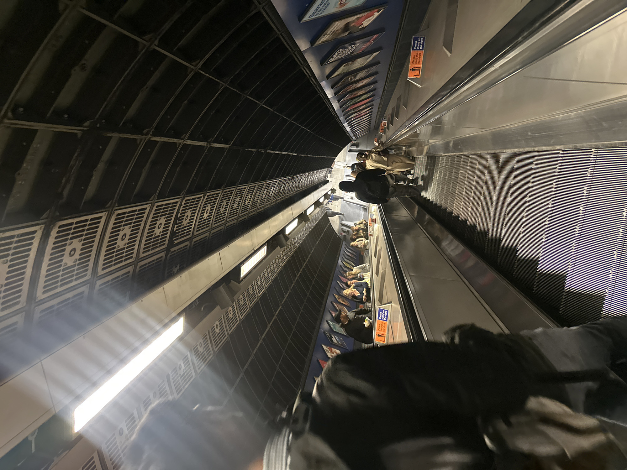 The image depicts a view looking down a long escalator within the London Underground system. The escalator has a metallic surface with ribbed steps. On either side of the escalator, there are handrails and signage. Orange signs with pictograms of a person and text such as "Please hold the handrail" and "Please stand on the right" are visible. The walls of the tunnel are lined with blue panels displaying advertisements and posters. The ceiling of the tunnel is curved and dark, with a grid-like structure and fluorescent lighting fixtures running along its length. Several people are visible on the escalator, some walking and others standing. The perspective is from slightly above and to the side, looking down the length of the escalator. The lighting is artificial and somewhat dim, characteristic of an underground station.