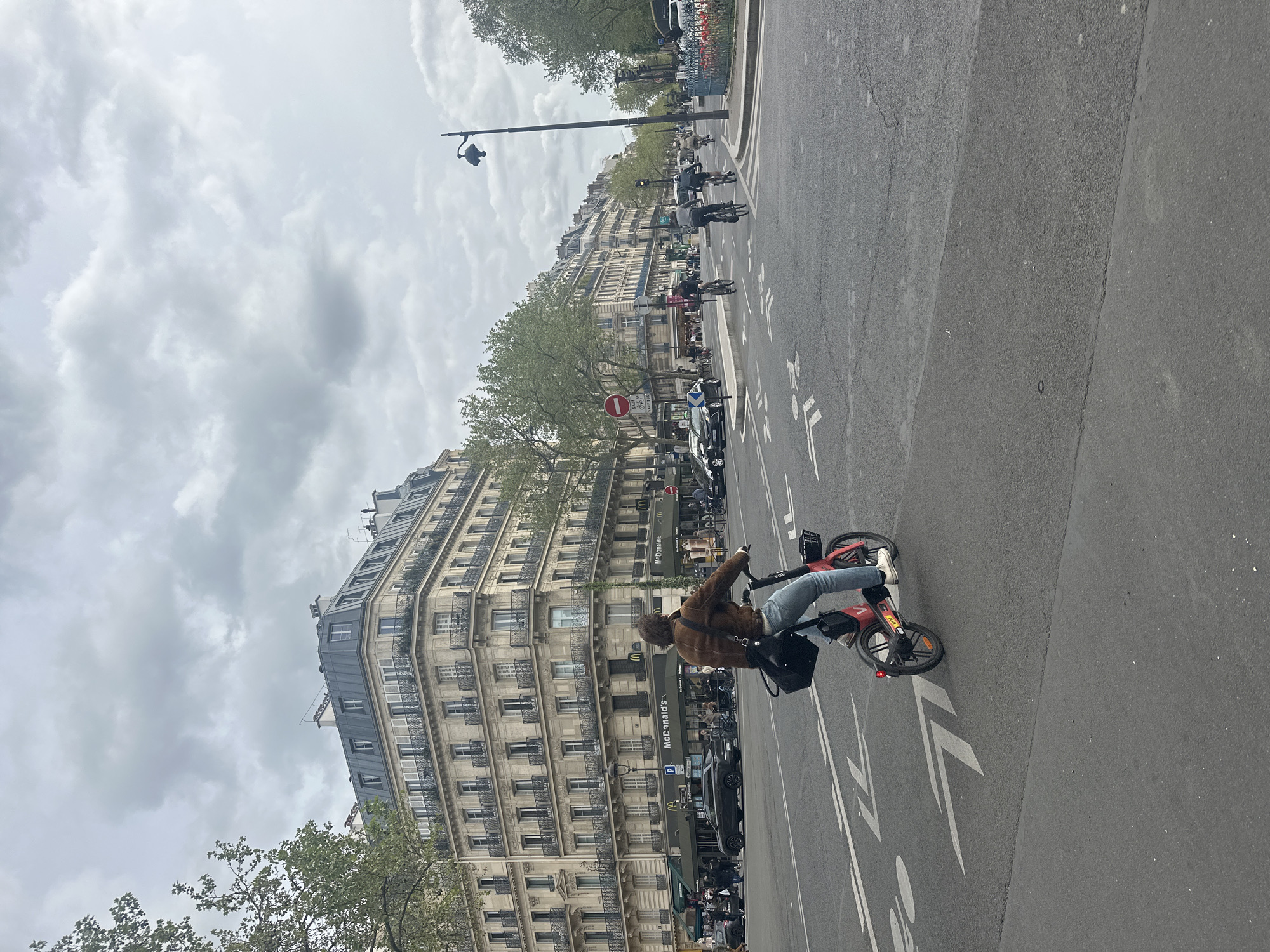 The image shows a street scene in Paris, France. A person wearing a brown jacket, blue jeans, and white shoes is riding a red Vélib' bike on a gray asphalt road marked with white directional arrows. The cyclist is positioned in the lower right corner of the frame. Behind the cyclist is a large building with multiple stories, many windows, and balconies. A "McDonald's" sign is visible on the building. A tree with green leaves is on the left edge of the frame. In the background are other people, cars, and bicycles on the street. The sky is overcast with gray clouds. A tall lamp post is visible in the middle of the image.