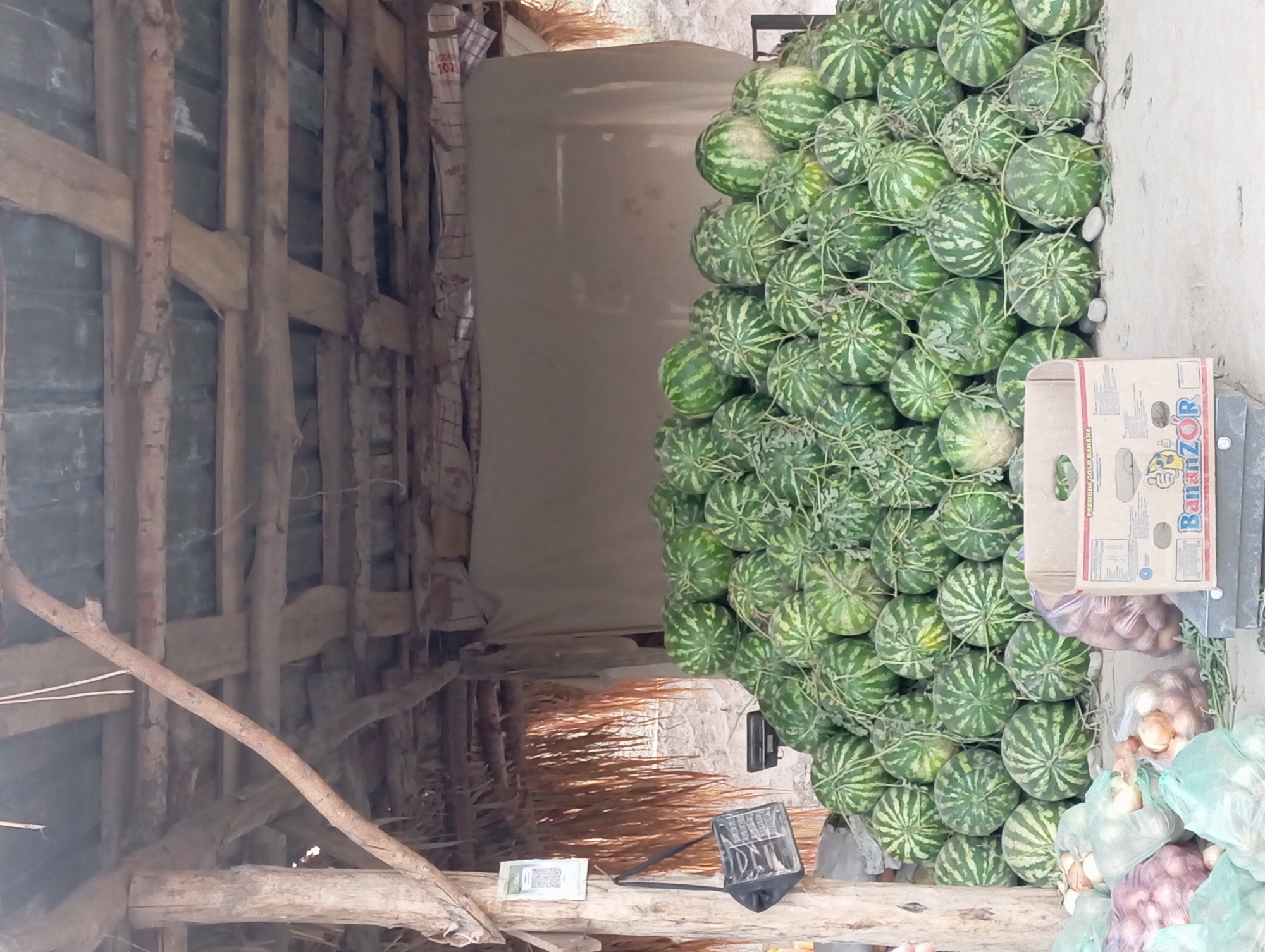 In the city of Садырбай, Kyrgyzstan, a stall displays a large pile of watermelons under a rustic, open-air shelter. The shelter's roof is constructed from corrugated metal and wooden beams, supported by rough-hewn wooden posts. To the left of the watermelons, a black bag hangs from a post, next to a sign with a QR code. In the foreground, several sacks of onions are visible, along with a cardboard box labeled "BananzOR". The ground is a sandy or dusty surface. No people are visible in the image. The overall scene depicts a market stall selling produce.