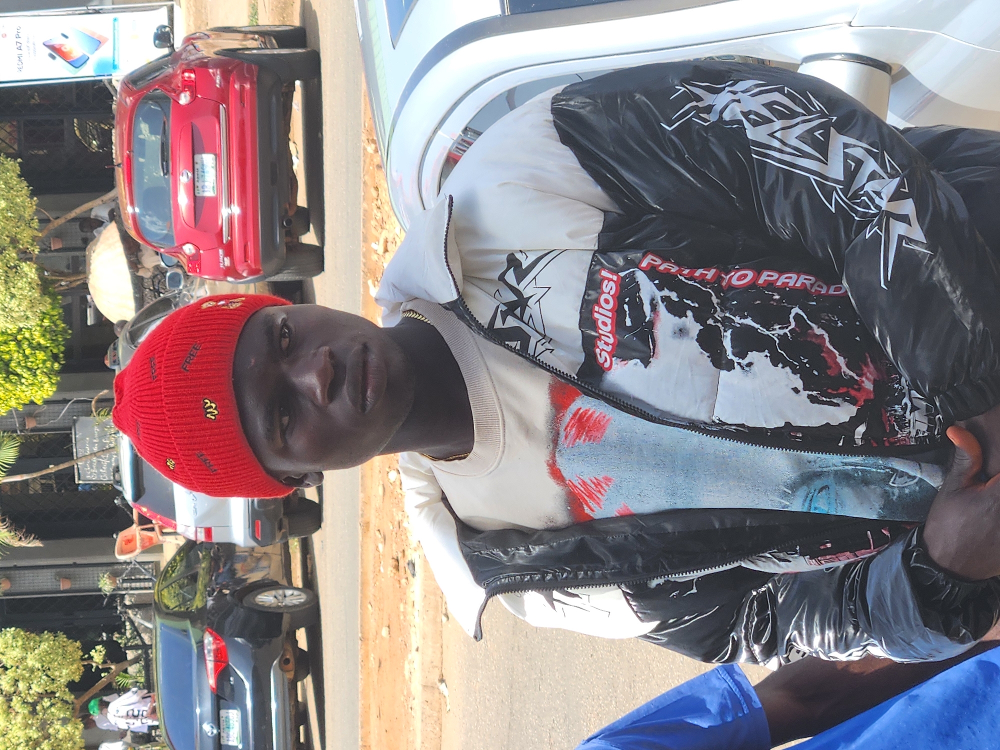 A waist-up shot depicts a dark-skinned man in a red beanie with yellow designs, leaning against a light-colored car. He wears a black and white jacket with text that reads "Studios! PATH YO PARADISE" over a white t-shirt with red and blue details. The location appears to be an urban street, with multiple cars of varying colors parked along the curb. A sign hangs in the background near a red car.