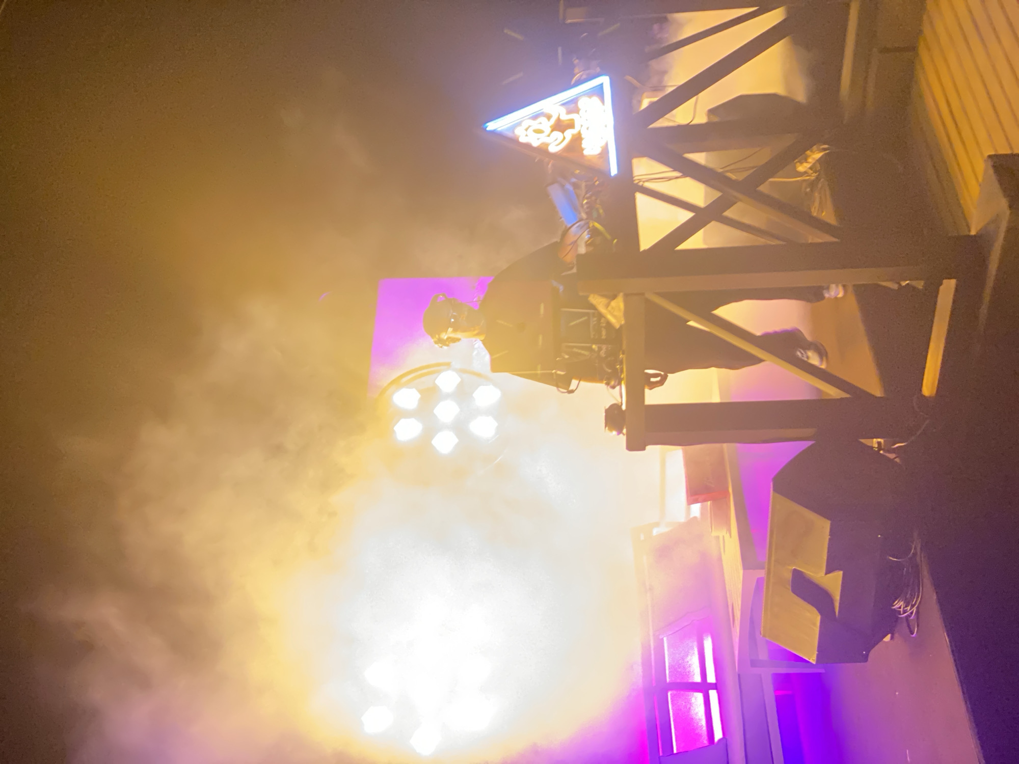The image depicts a DJ performing on a stage, likely during an outdoor event in Taghazout, Morocco. The DJ, wearing a headset and sunglasses, stands behind a DJ console adorned with a triangular neon sign featuring a cartoon character. The stage is illuminated by bright, hazy lights, with purple and yellow hues dominating the scene, suggesting a vibrant, possibly nighttime, atmosphere. A large speaker is visible in the foreground. The background features a simple white couch and table setup, possibly for VIP guests or performers. The overall impression is of a lively music performance in a coastal town, characteristic of Taghazout's entertainment scene.