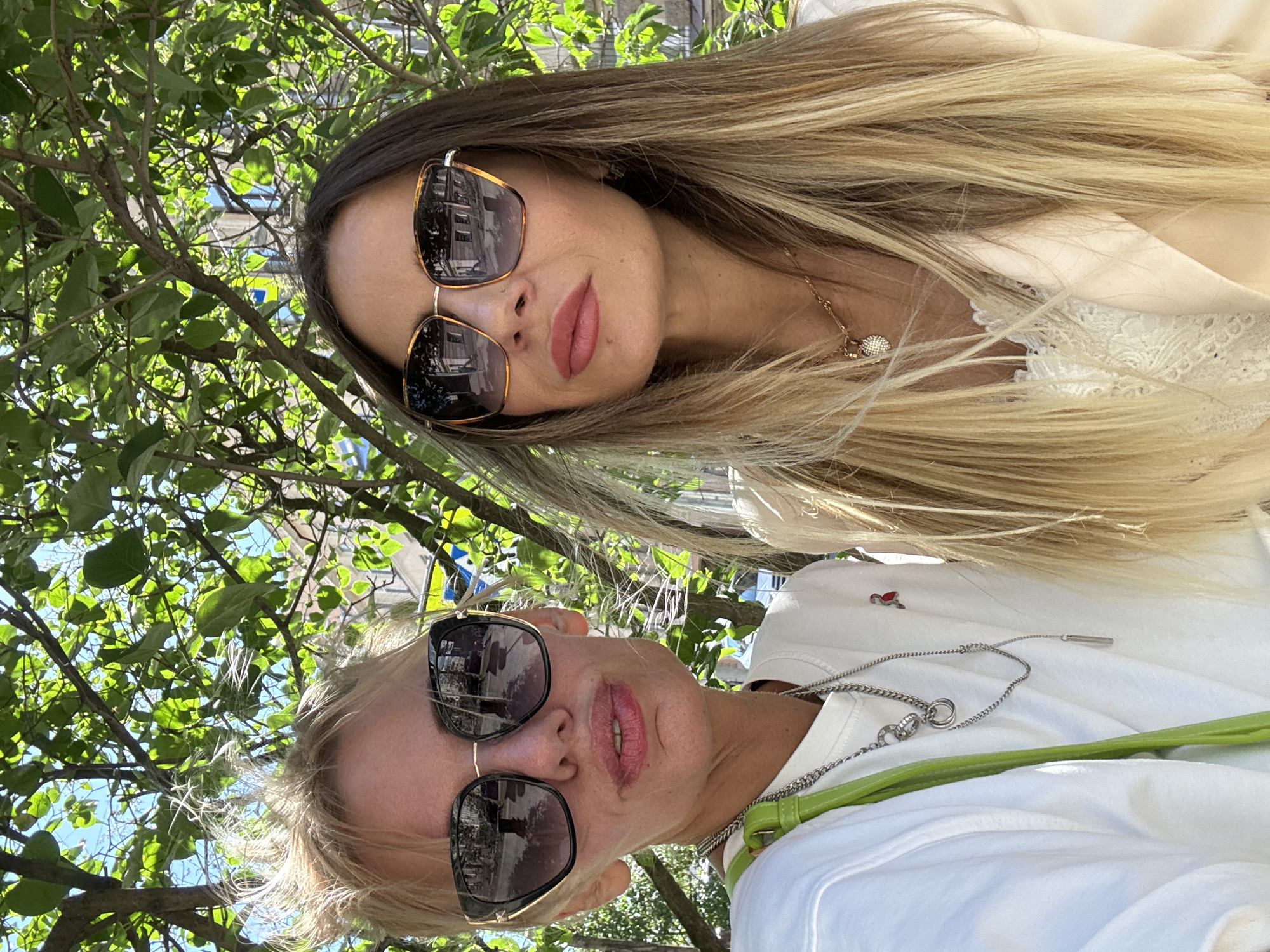 Two women are taking a selfie outdoors. The woman in the foreground has blonde hair and is wearing sunglasses and a white shirt. The woman behind her has long blonde and brown hair and is wearing sunglasses and a light-colored top. Both women are wearing necklaces. They are sitting under a tree with green leaves, and buildings are visible in the background. The sun is shining, suggesting it is daytime and the weather is pleasant. The overall mood appears relaxed and casual, possibly indicating a leisurely outing. No visible text is present in the image.