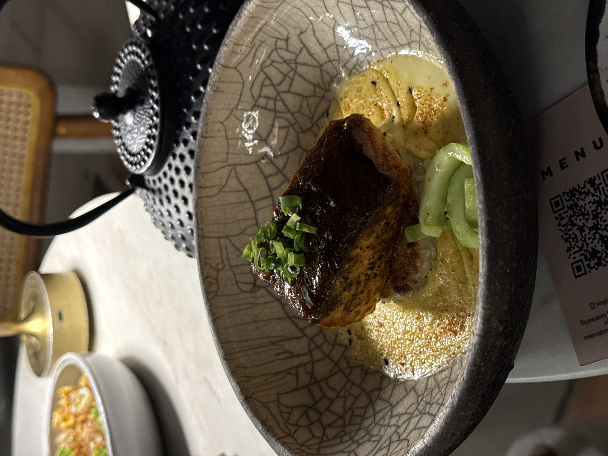 The image depicts a plate of food on a table, likely in a restaurant. The plate contains what appears to be a piece of glazed meat, garnished with green onions, accompanied by a creamy sauce and some cucumber slices. The plate itself has a cracked, ceramic-like texture. To the left of the plate is a black teapot and in the lower left, another dish.
The image contains a partial view of a menu, referencing "Bluewaters Island", which is a location in دبي, United Arab Emirates.